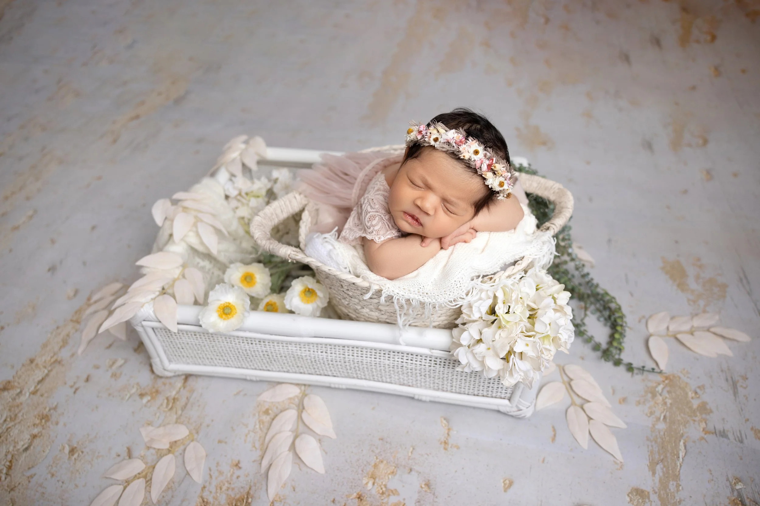 A sleeping baby girl with a floral headband, lying in a white woven basket surrounded by white and yellow flowers and light-colored leaves on a textured gray surface.