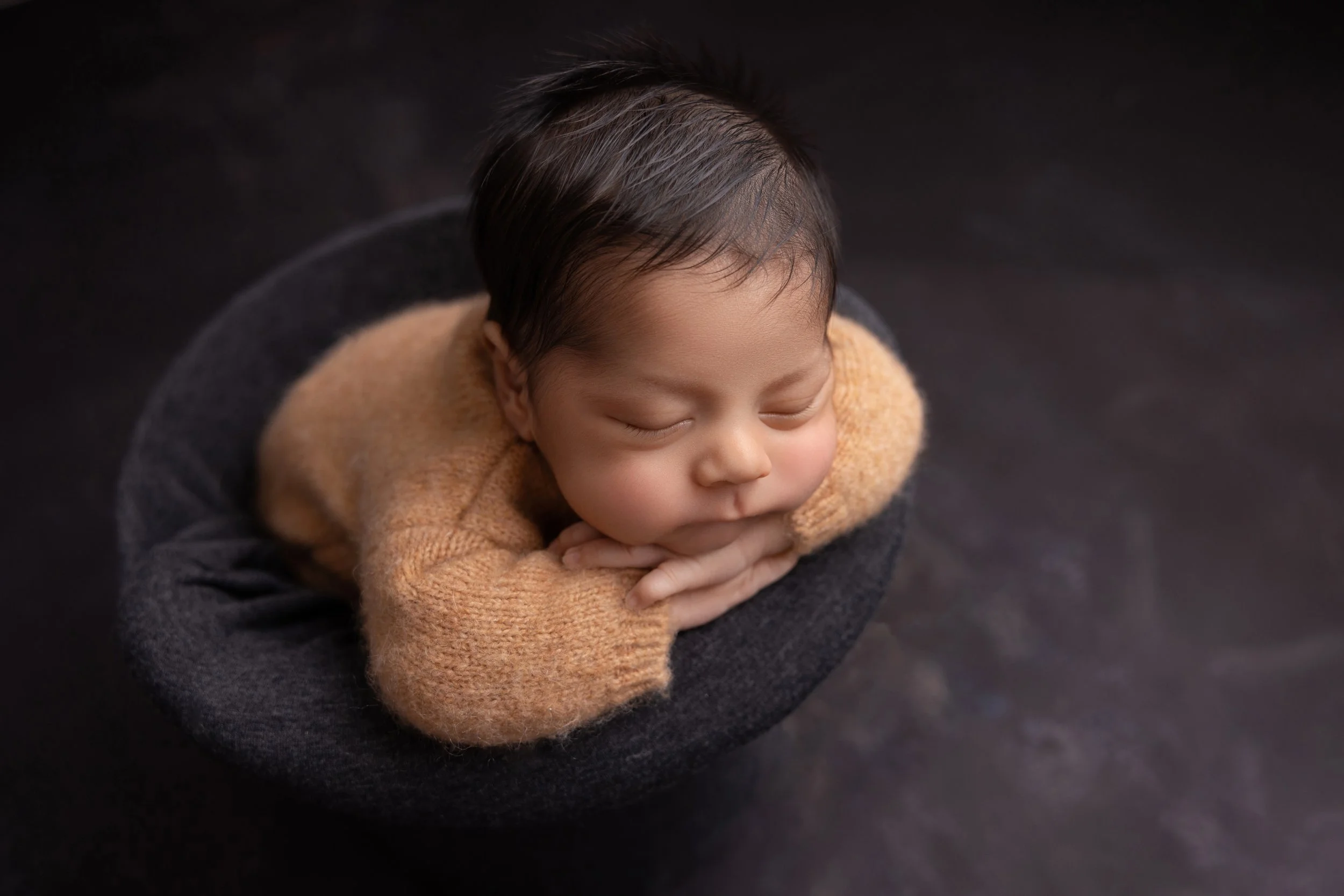 A sleeping baby with dark hair, resting their head on their folded hands, lying inside a dark gray woven basket against a dark background.