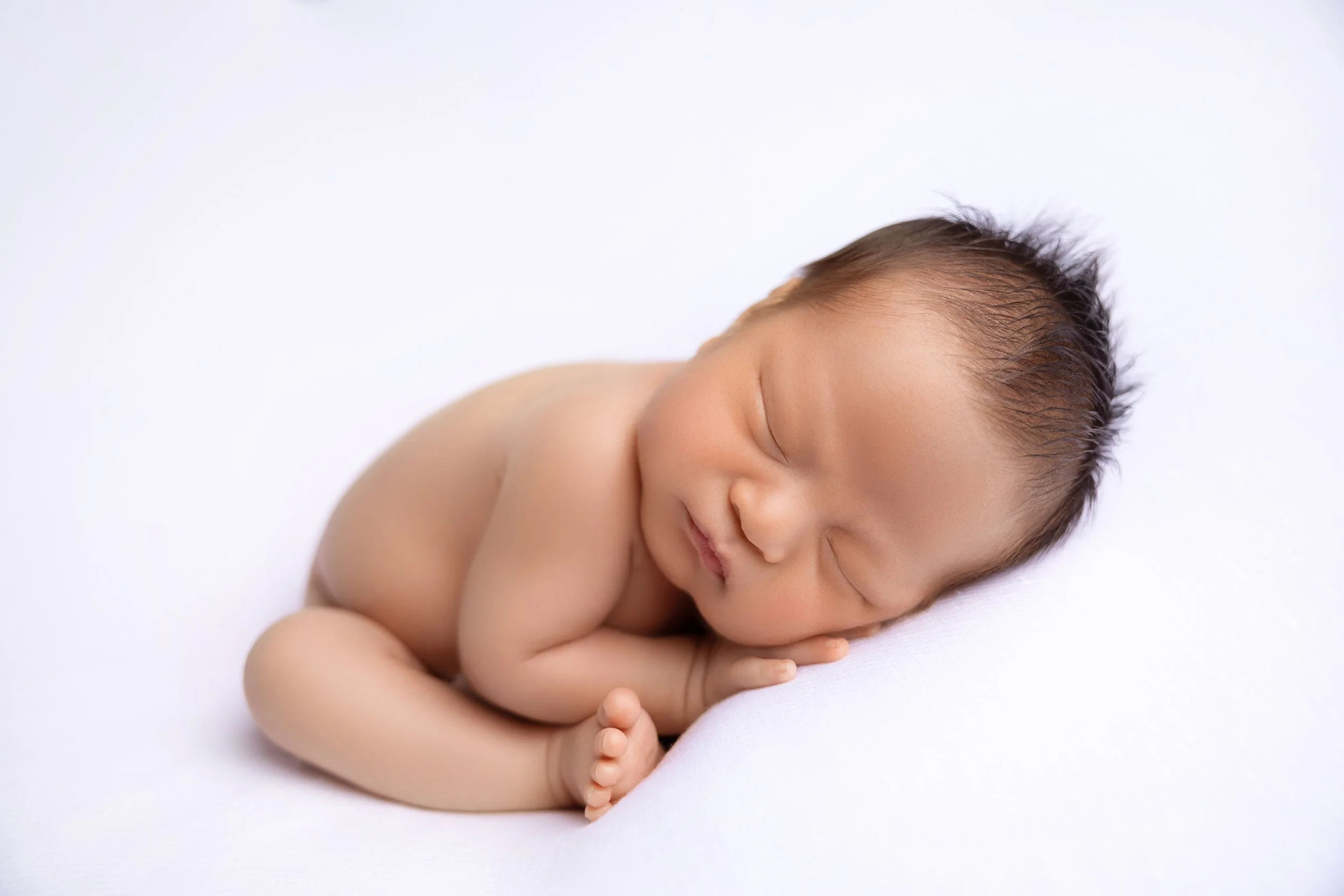 A sleeping newborn baby curled up on a white surface with a peaceful expression.