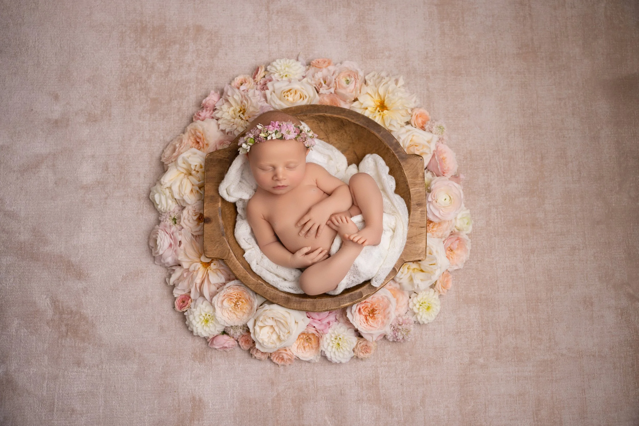 A sleeping newborn baby lying in a wooden bowl, surrounded by pink and white flowers on a soft pink background.
