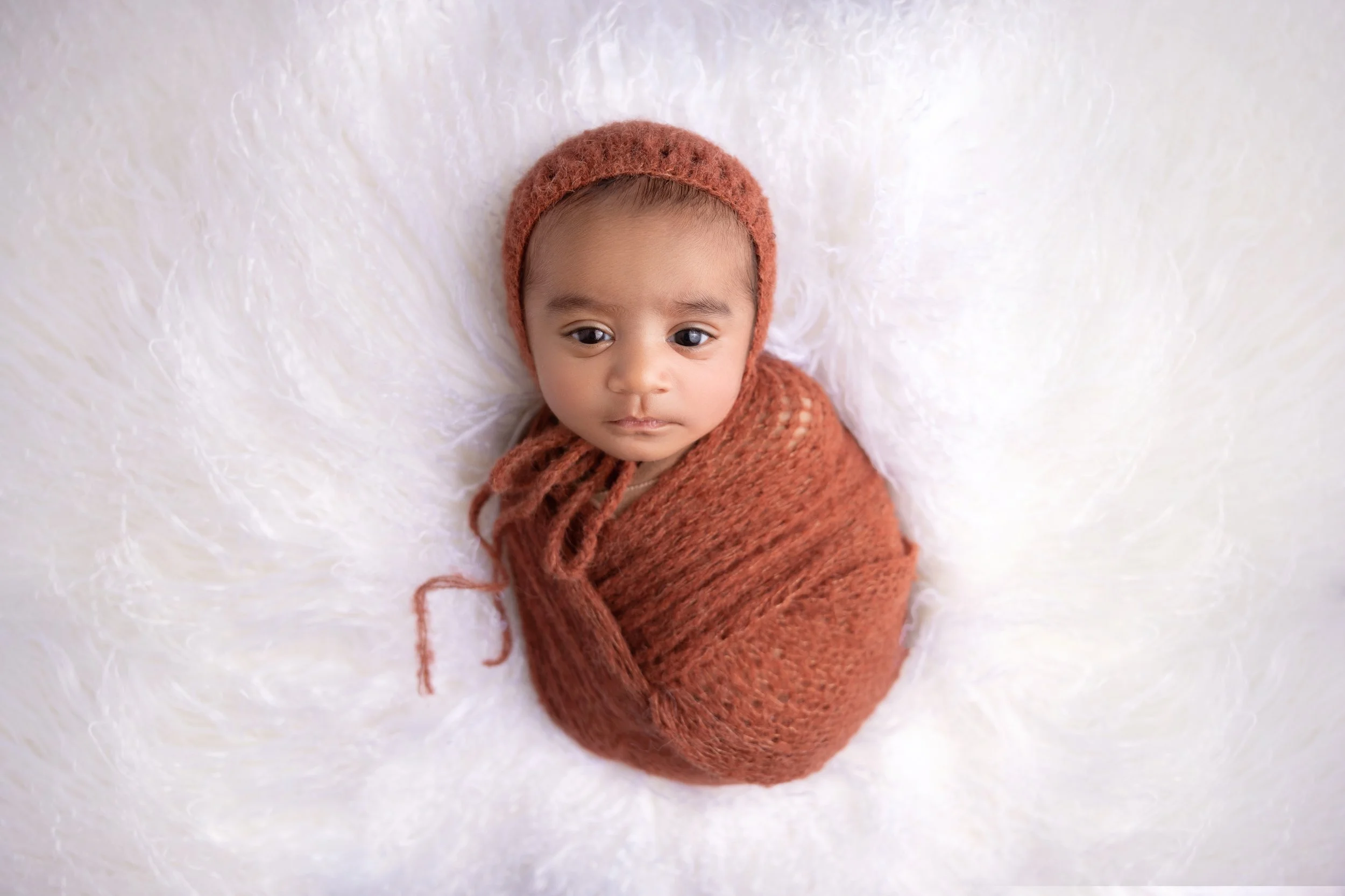 A young child wrapped in a brown knitted blanket and wearing a matching brown headband, lying on a soft white blanket.