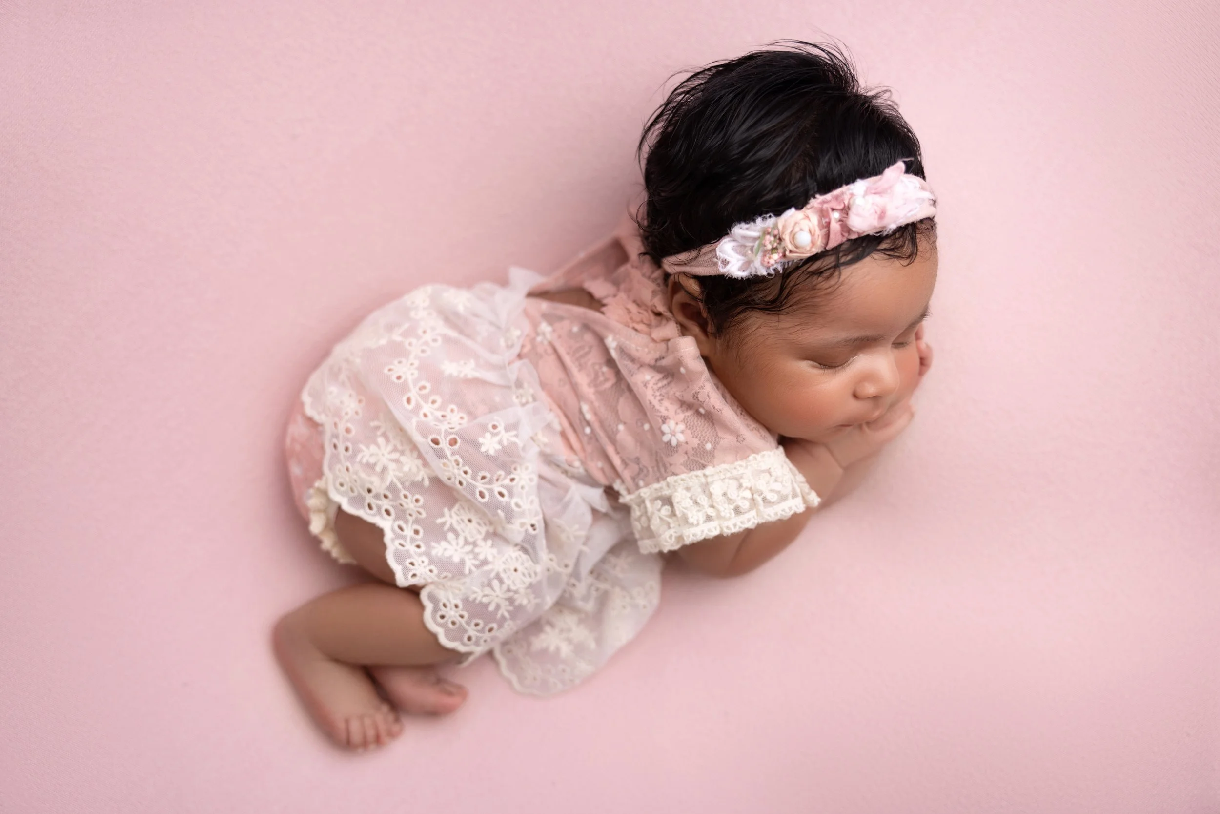 Young baby girl, dressed in a pink lace dress with floral embroidery, lying on a pink surface with a matching pink floral headband, sleeping peacefully.