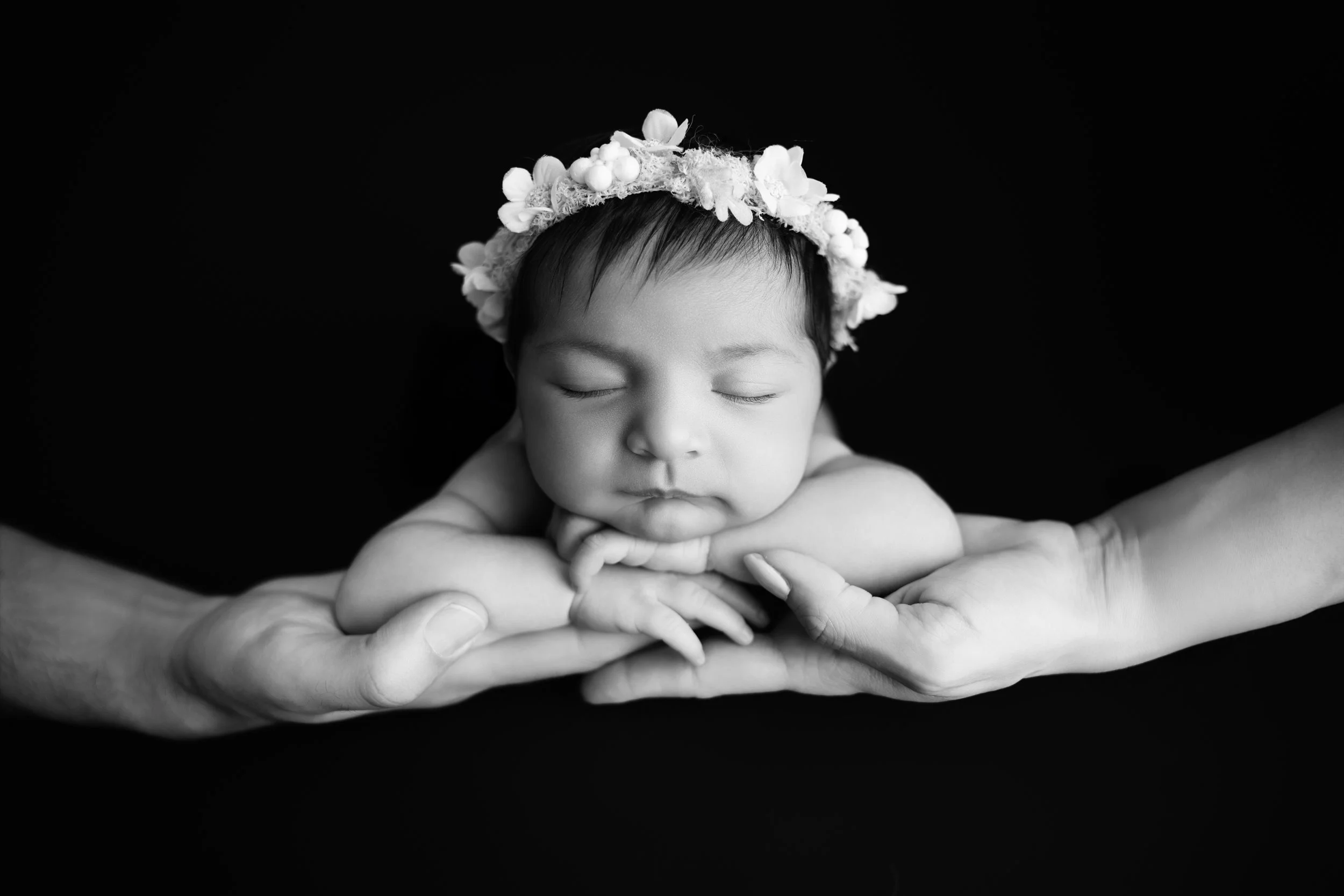 Black-and-white photo of a sleeping baby with a flower headband, resting on an adult's hands against a dark background.