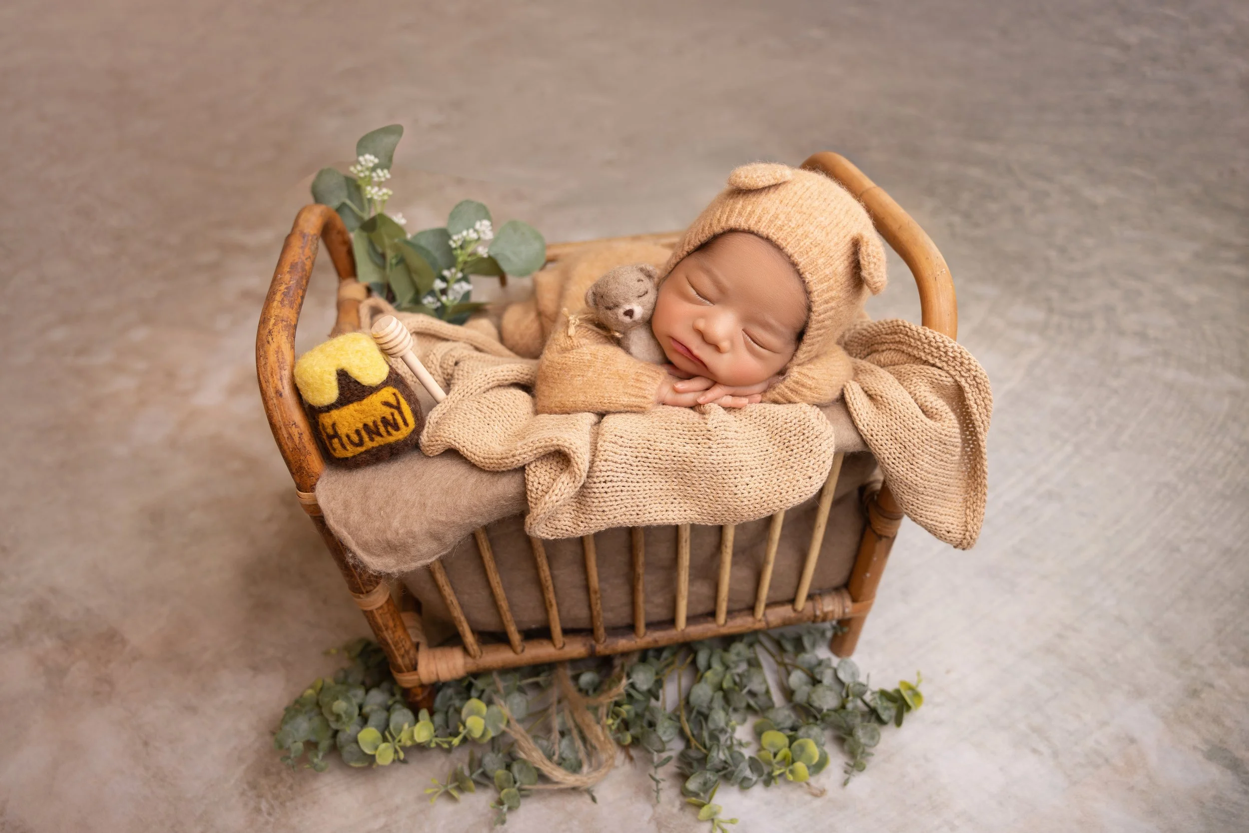 A sleeping baby dressed in a tan bear hat and blanket, lying in a vintage wicker bassinet with decorative green foliage underneath, and a small knitted honey pot with the word 'Hunny' beside him.