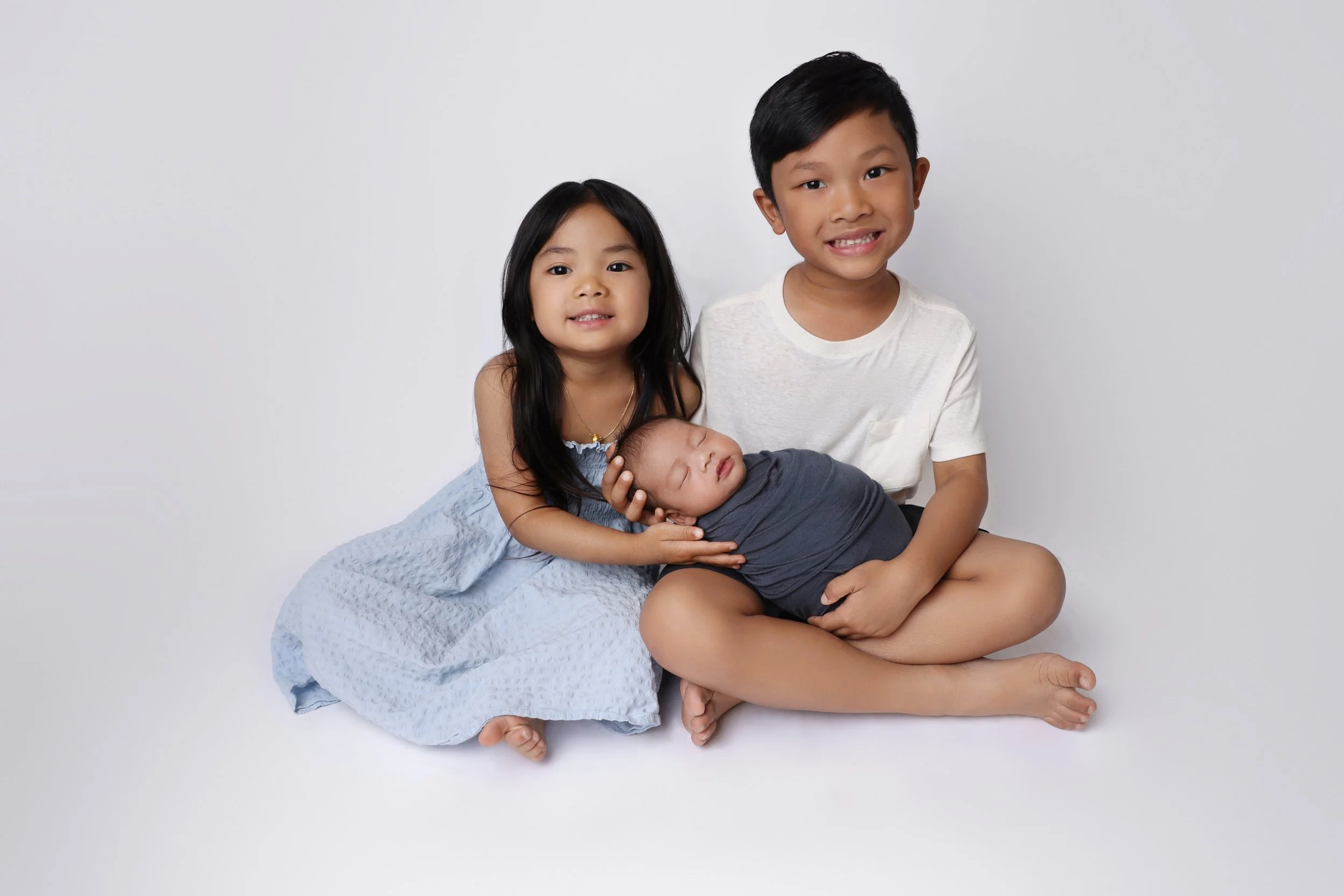 Three children, two standing and one sleeping, sitting on a white background. The girl on the left has long dark hair and wears a blue dress; the boy on the right has short dark hair and wears a white shirt; the baby in the middle is sleeping and wra