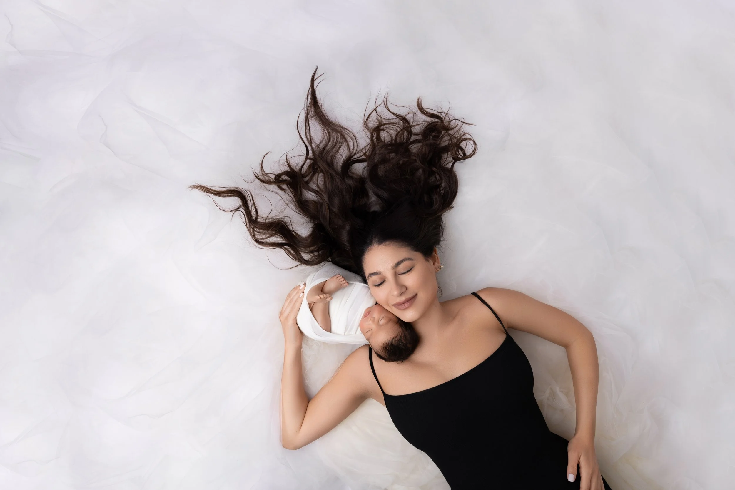 A woman with long, dark hair lying on a white surface, holding a sleeping baby wrapped in a white blanket. The woman is wearing a black spaghetti strap top, and she has a peaceful expression with her eyes closed.