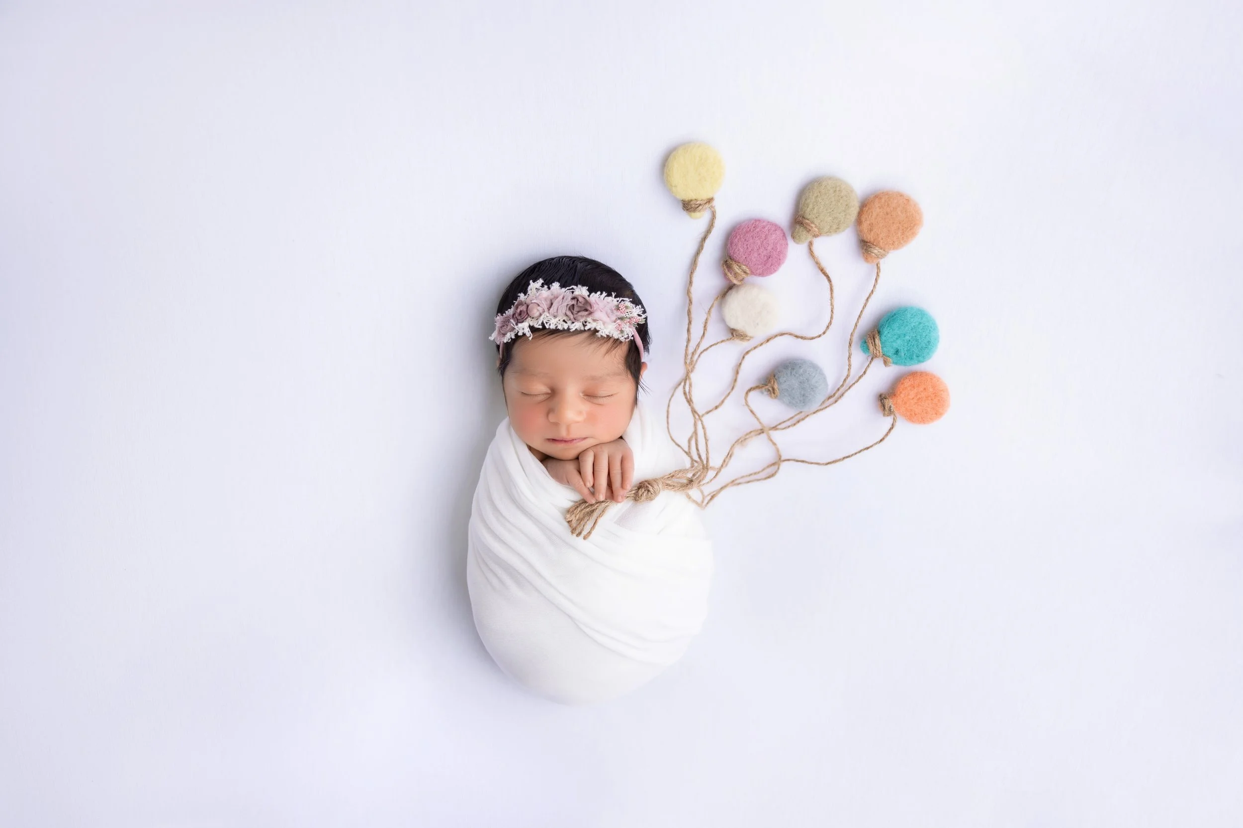Newborn baby girl wrapped in a white swaddle, lying on a white background, with multi-colored felt balloons attached to strings near her head.