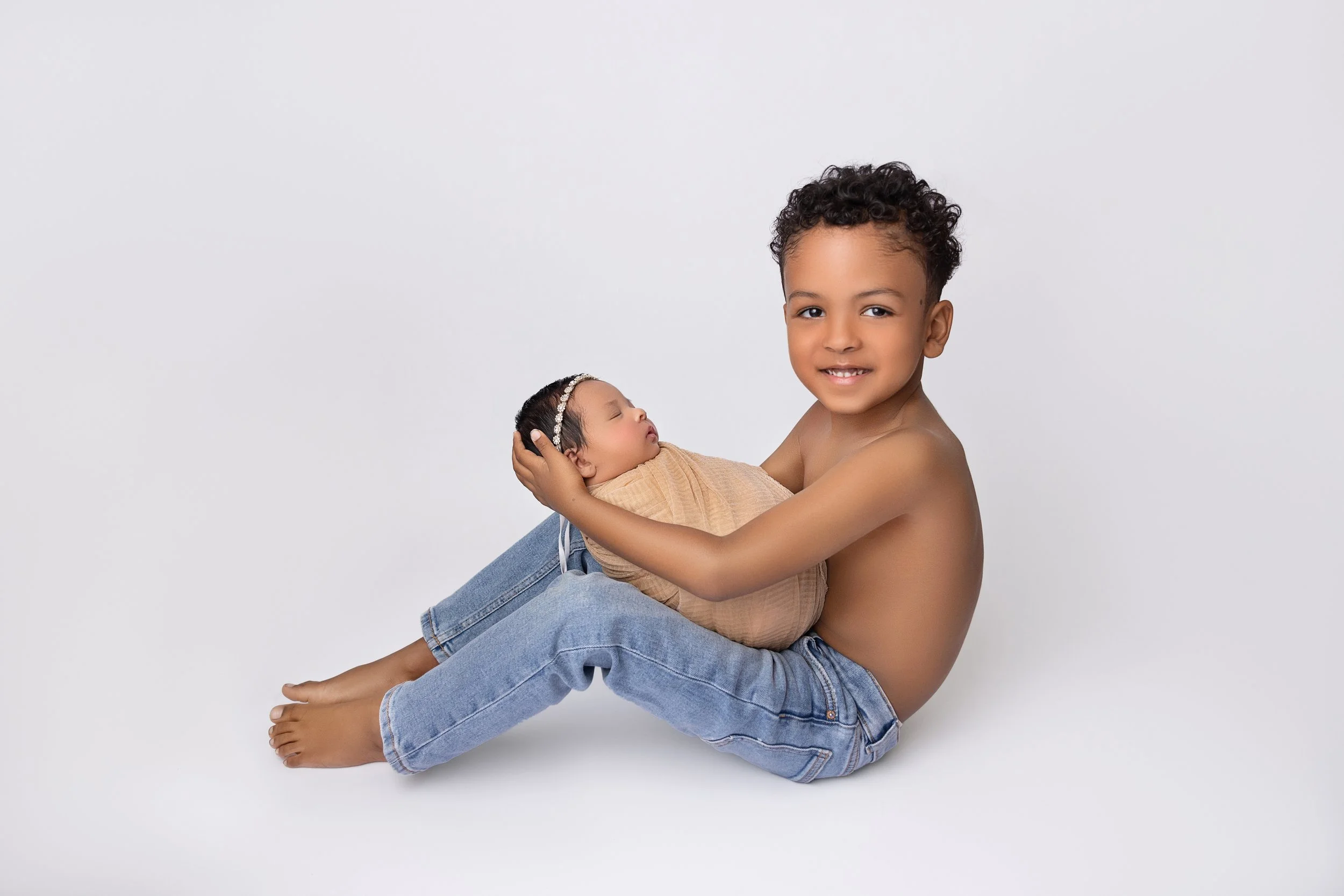 A young boy with curly hair, shirtless, holding a sleeping newborn girl wrapped in a beige blanket, against a plain white background.