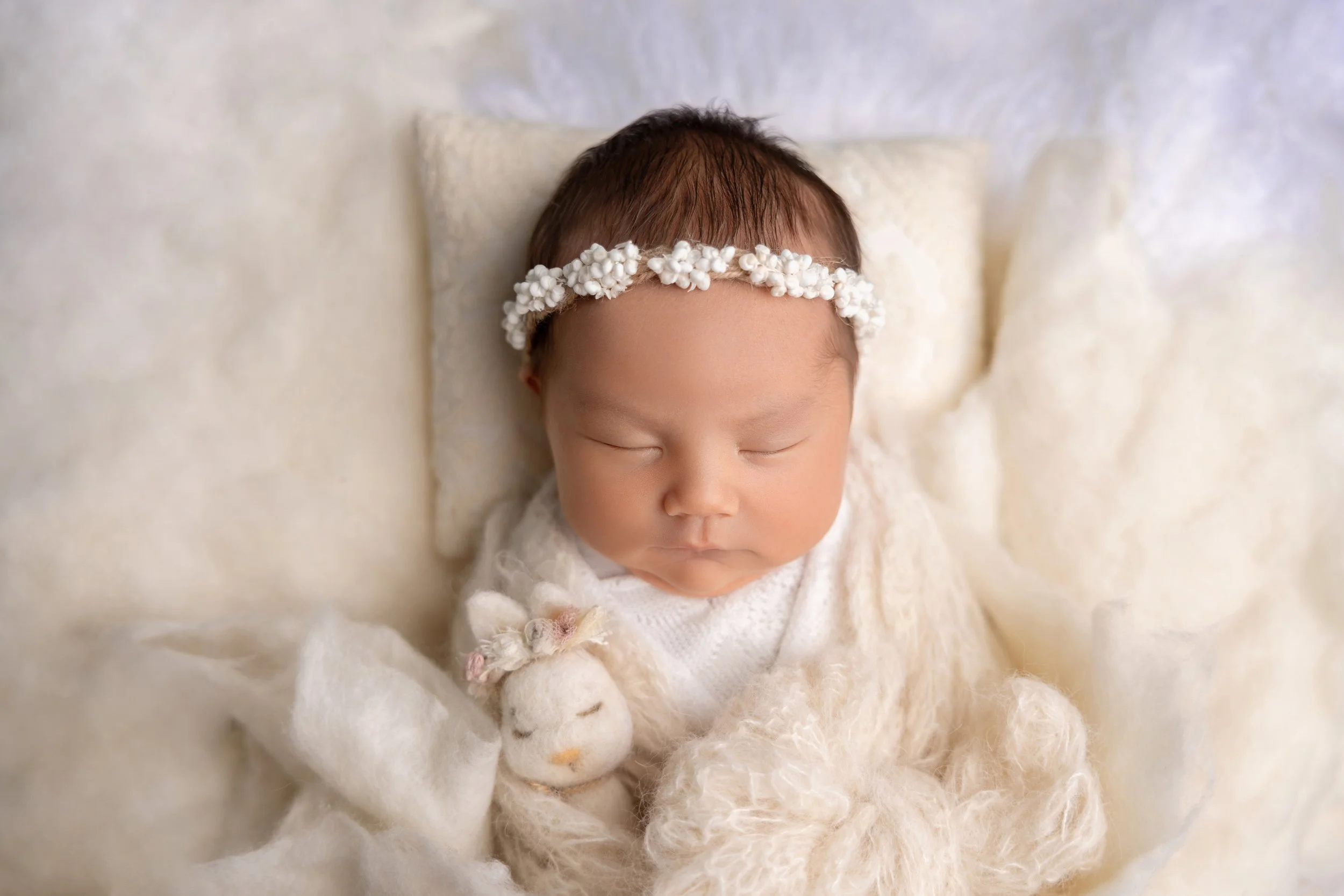 A sleeping newborn baby with a floral headband, lying on a cream-colored blanket with a plush lamb toy.