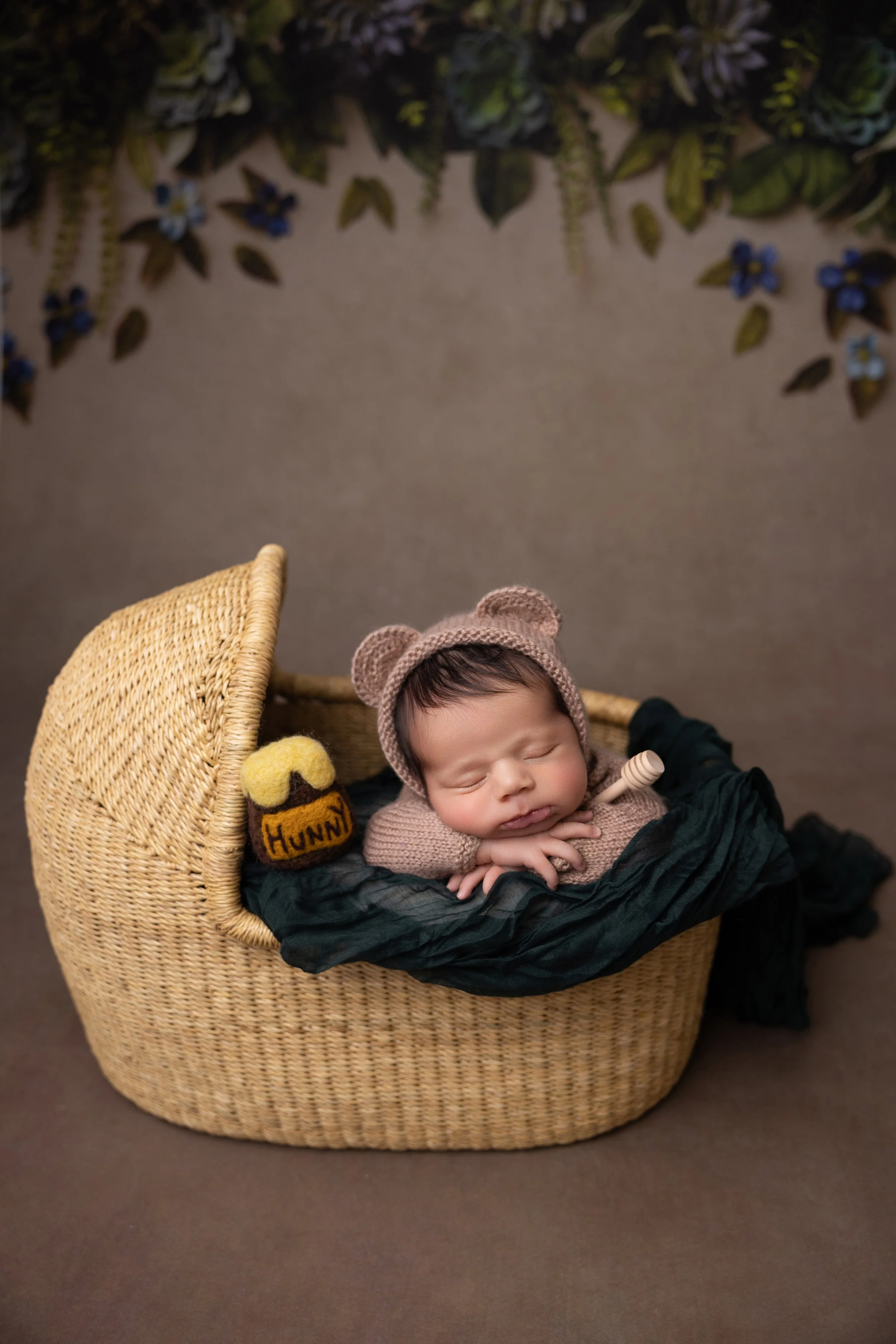 A sleeping baby dressed in a bear hat lies on its stomach on black fabric inside a woven basket, with a decorative sock that says "Hunny" beside it, against a backdrop of green foliage and blue flowers.