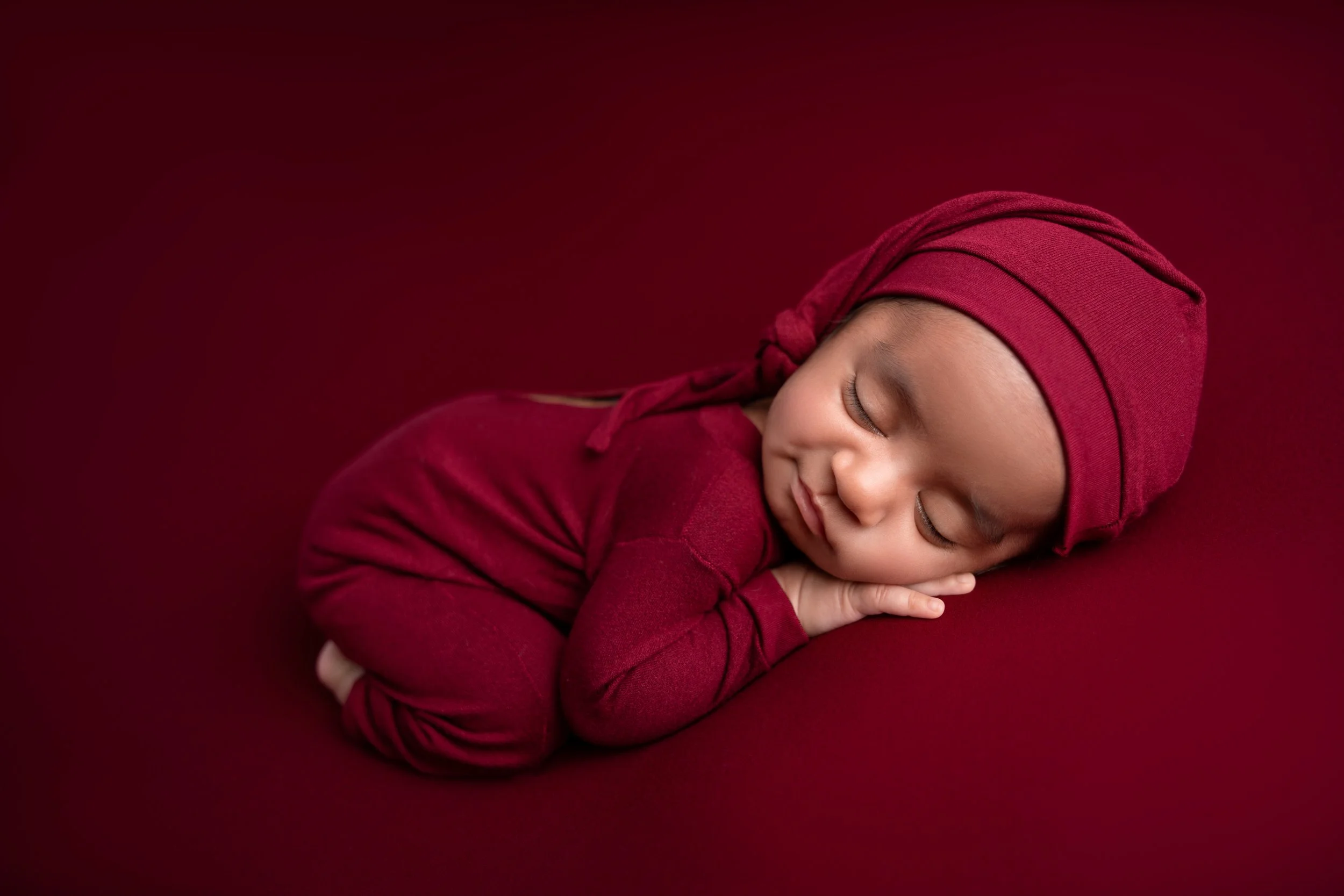 Young child sleeping peacefully on a maroon surface, wearing a maroon outfit and matching headscarf.
