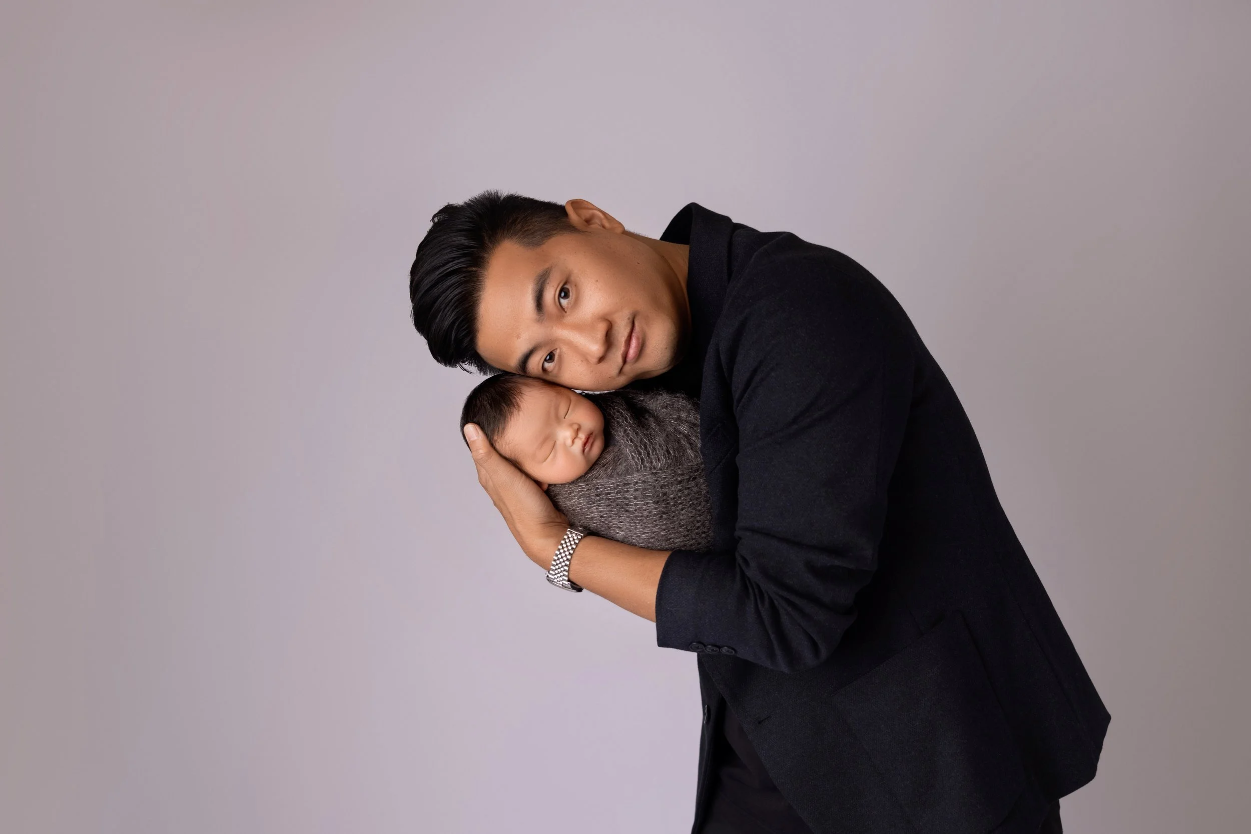 A man in a black blazer holding a sleeping baby wrapped in a gray blanket, against a plain gray background.