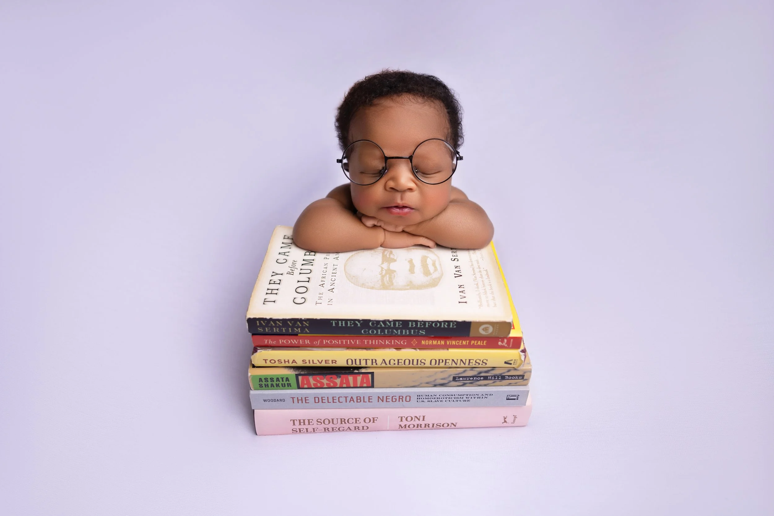 A baby with glasses resting its chin on a stack of eight books on a light purple background.