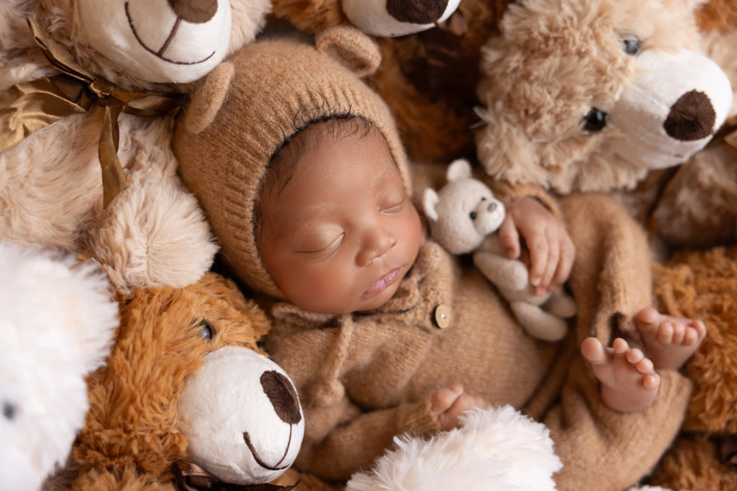 A sleeping baby bundled in a brown knit outfit and hat, surrounded by stuffed animal bears in various shades of brown and white.