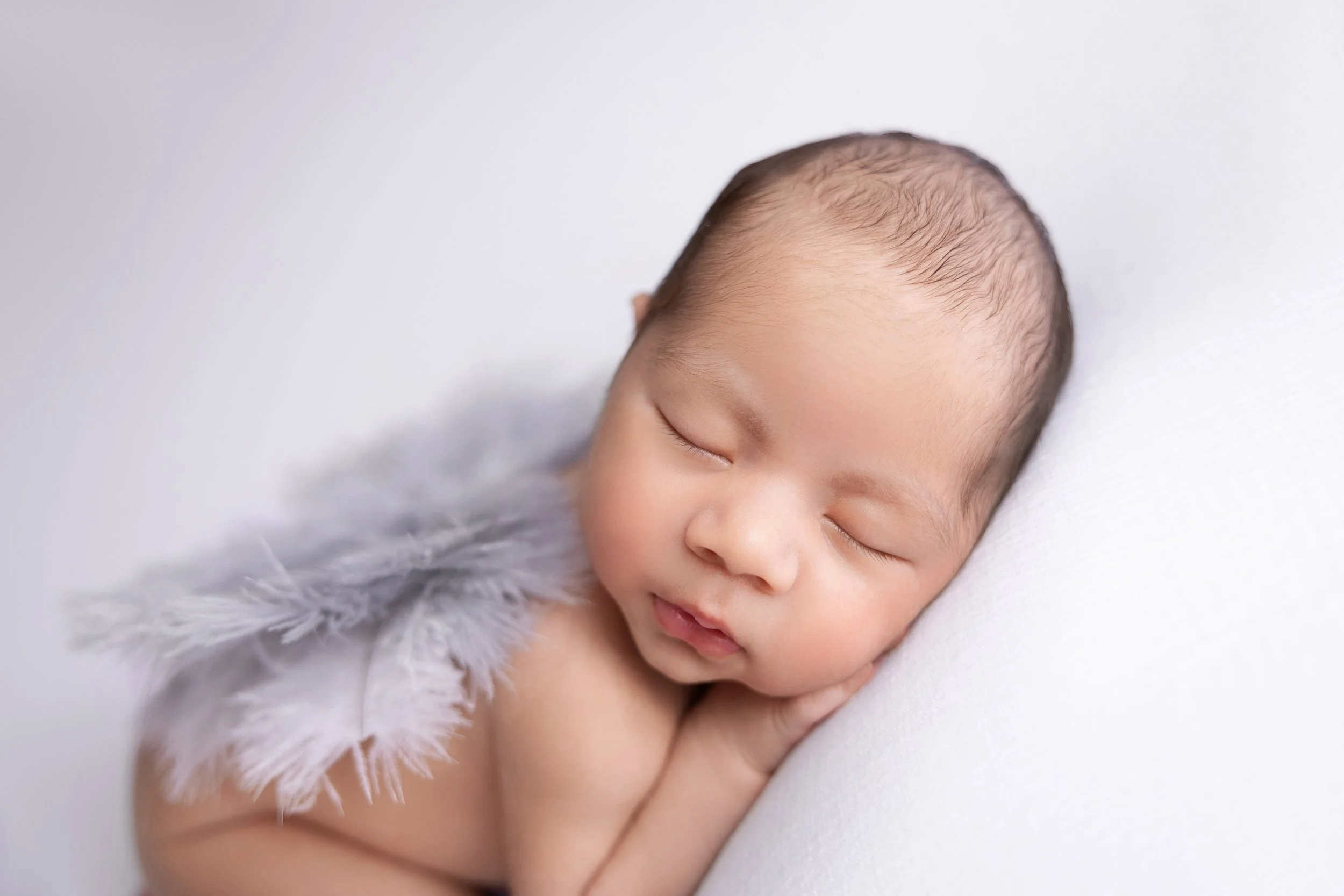 A sleeping baby resting on a white pillow, wearing a soft, fluffy gray feathered accessory.