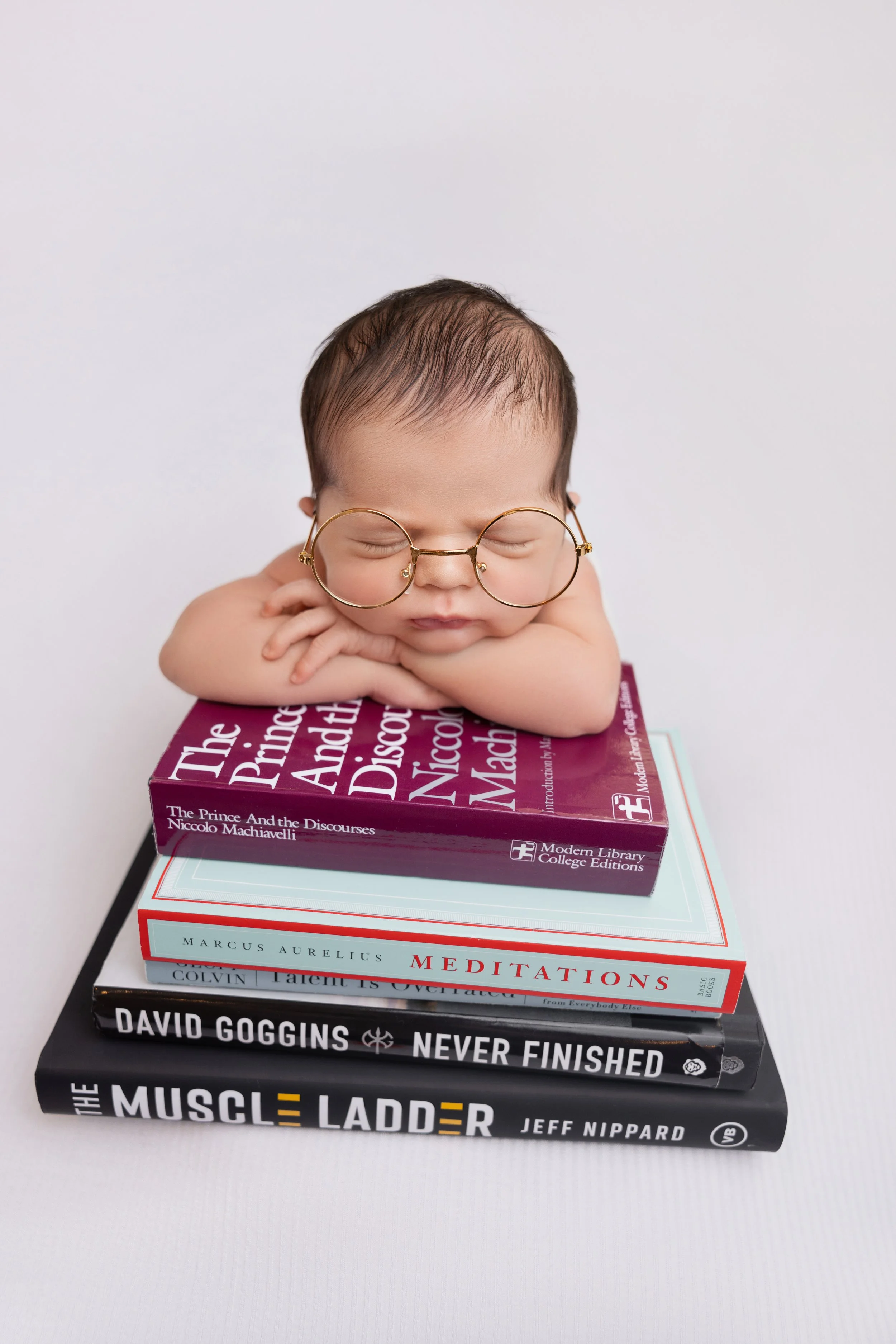 A baby with glasses resting their chin on folded arms atop a stack of books.