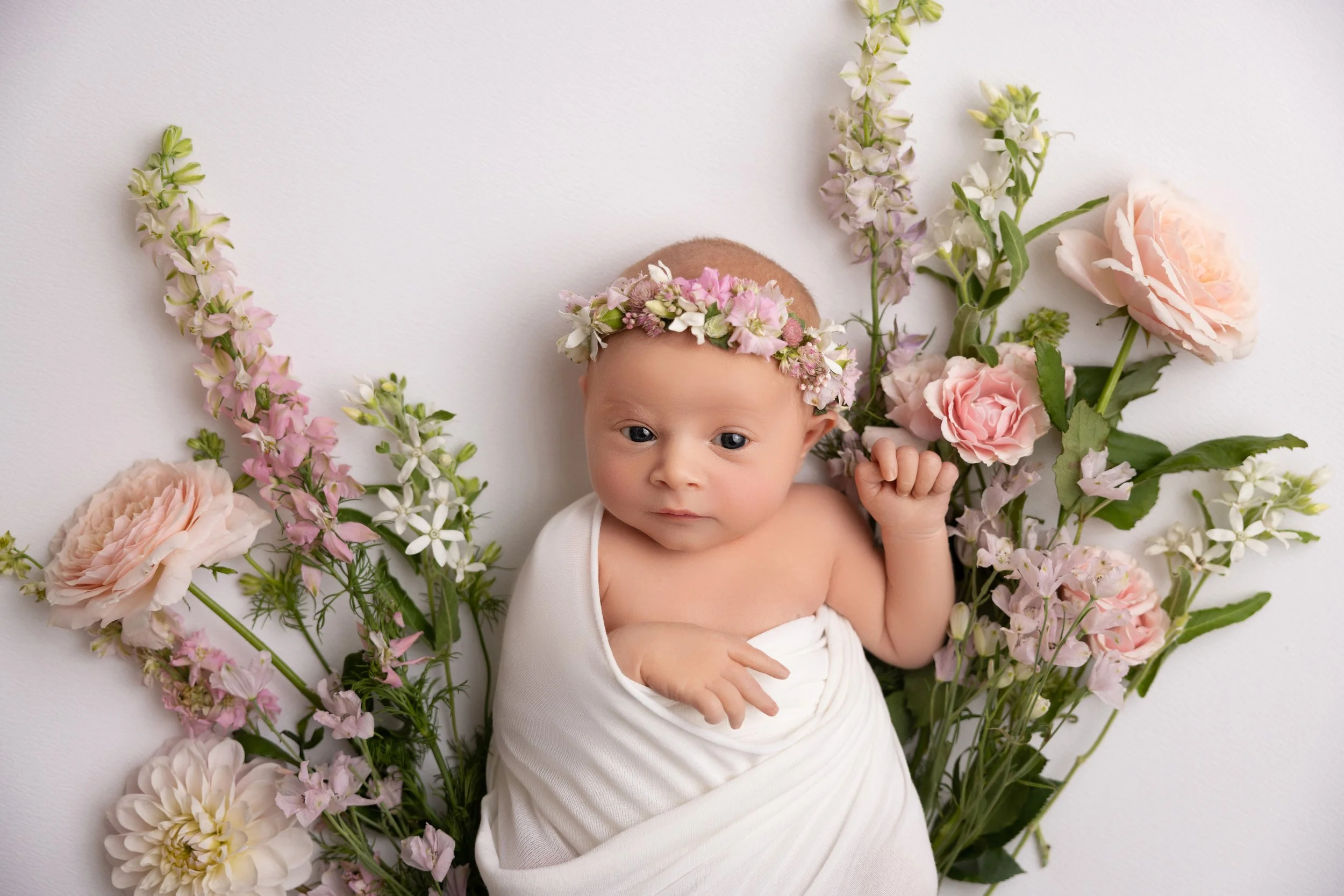A baby lying on a white surface surrounded by pink and white flowers, wearing a floral crown and wrapped in a white cloth.