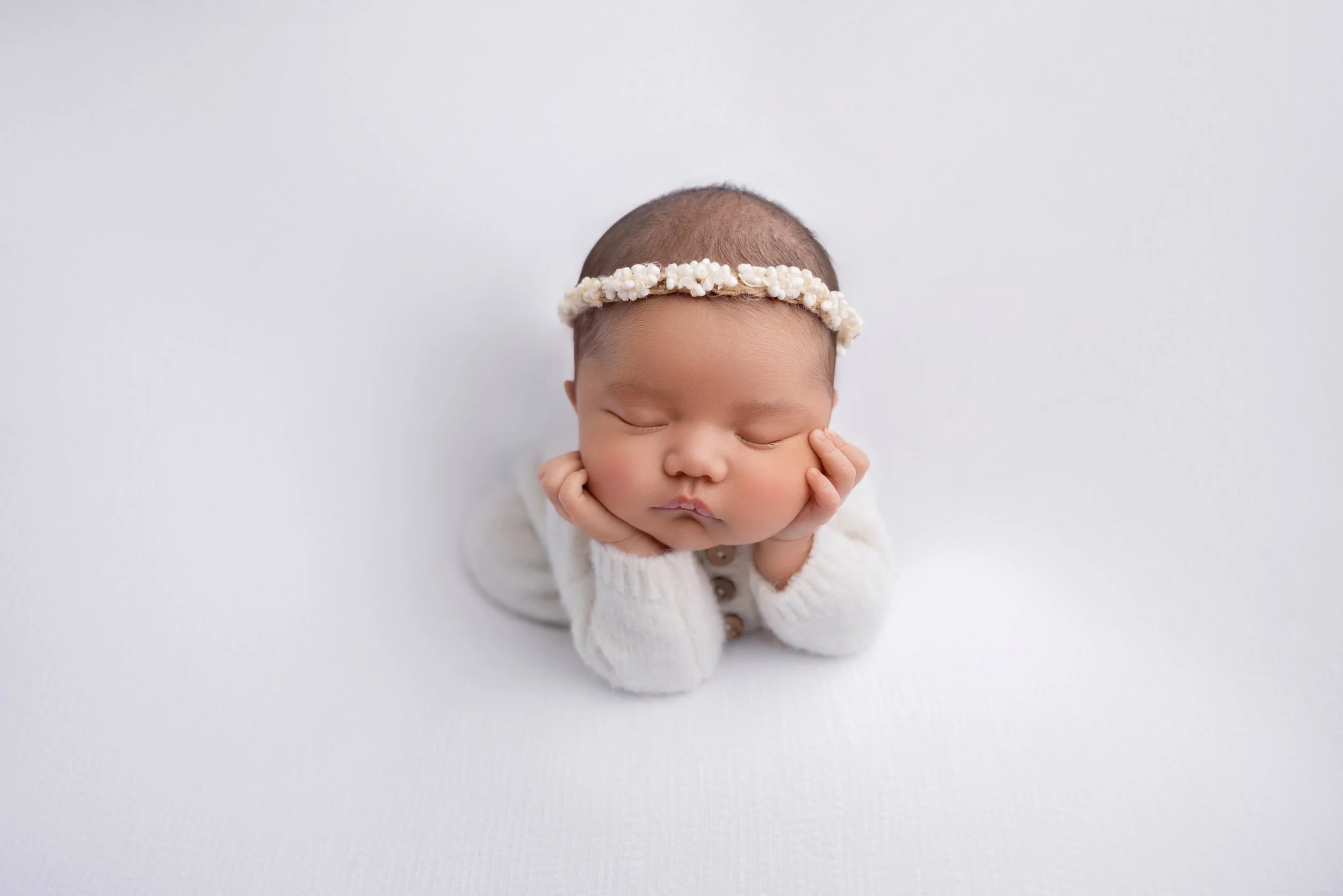 A baby with a floral headband, resting face down on a white surface, wearing a white knitted sweater.