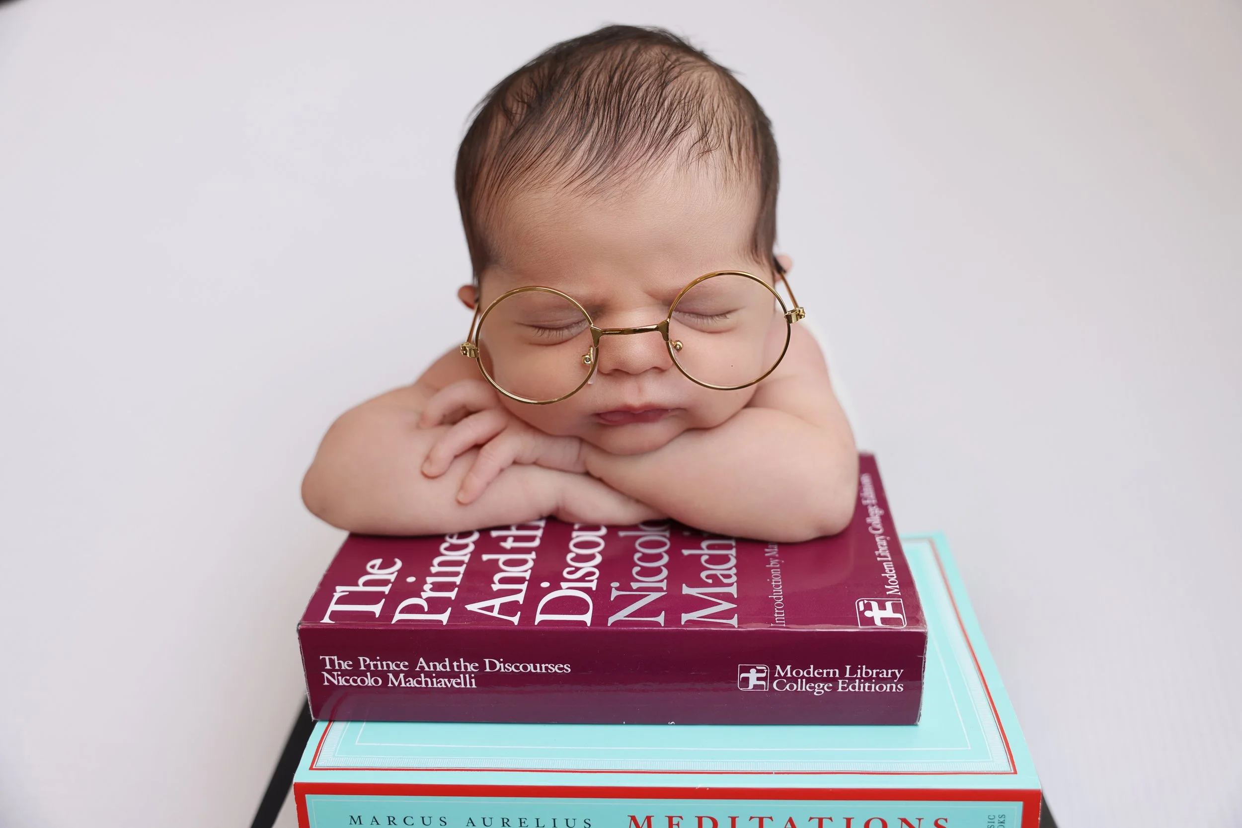 A baby with glasses resting its head on folded arms on top of two stacked books, one titled "The Prince and the Discourse" and the other titled "Meditations".