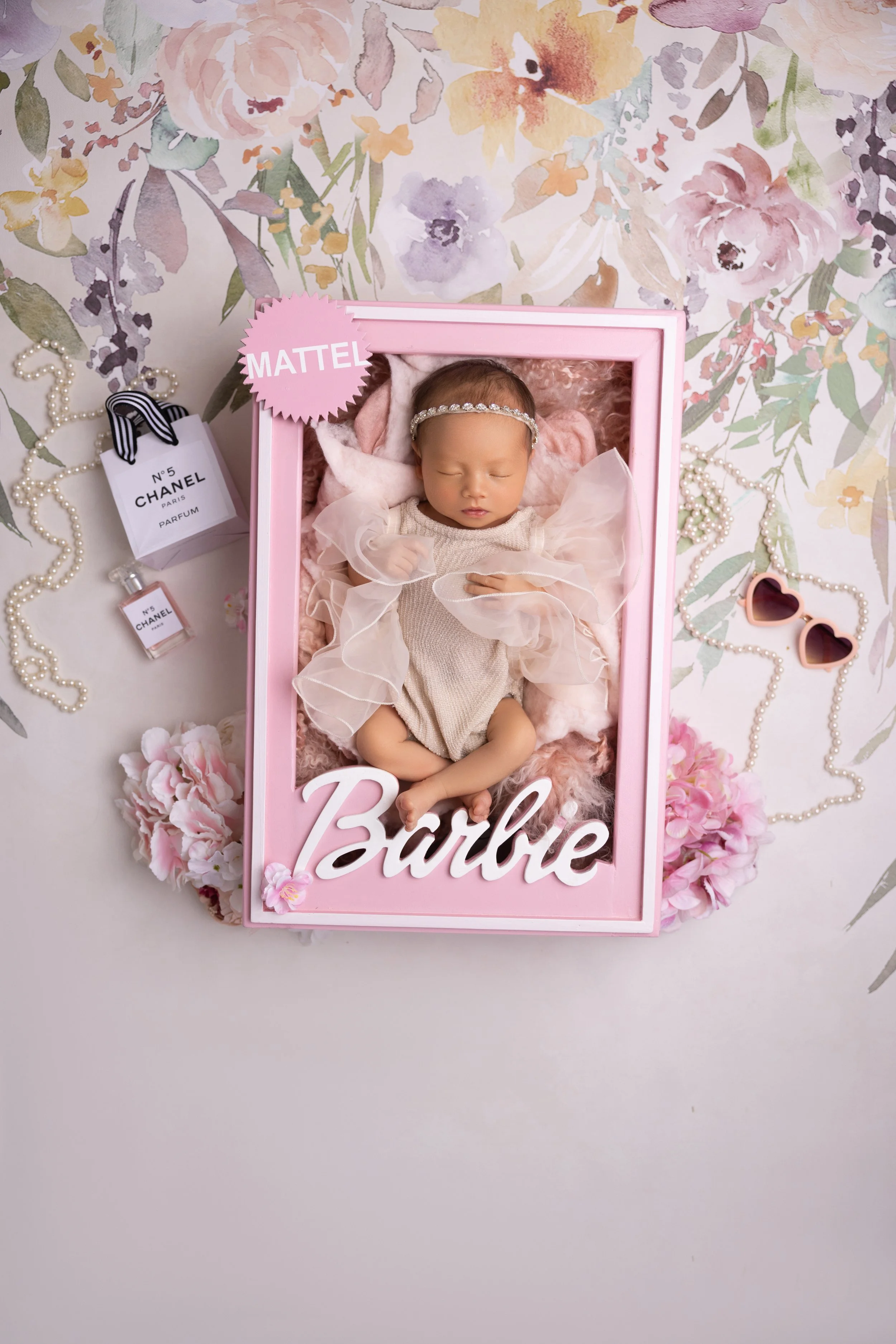 A sleeping baby doll inside a pink box with the name 'Barbie' on it. The box has a pink frame and is decorated with artificial pink flowers around the base. The baby doll wears a beige dress with sheer pink ruffles and a headband, lying on pink fluff