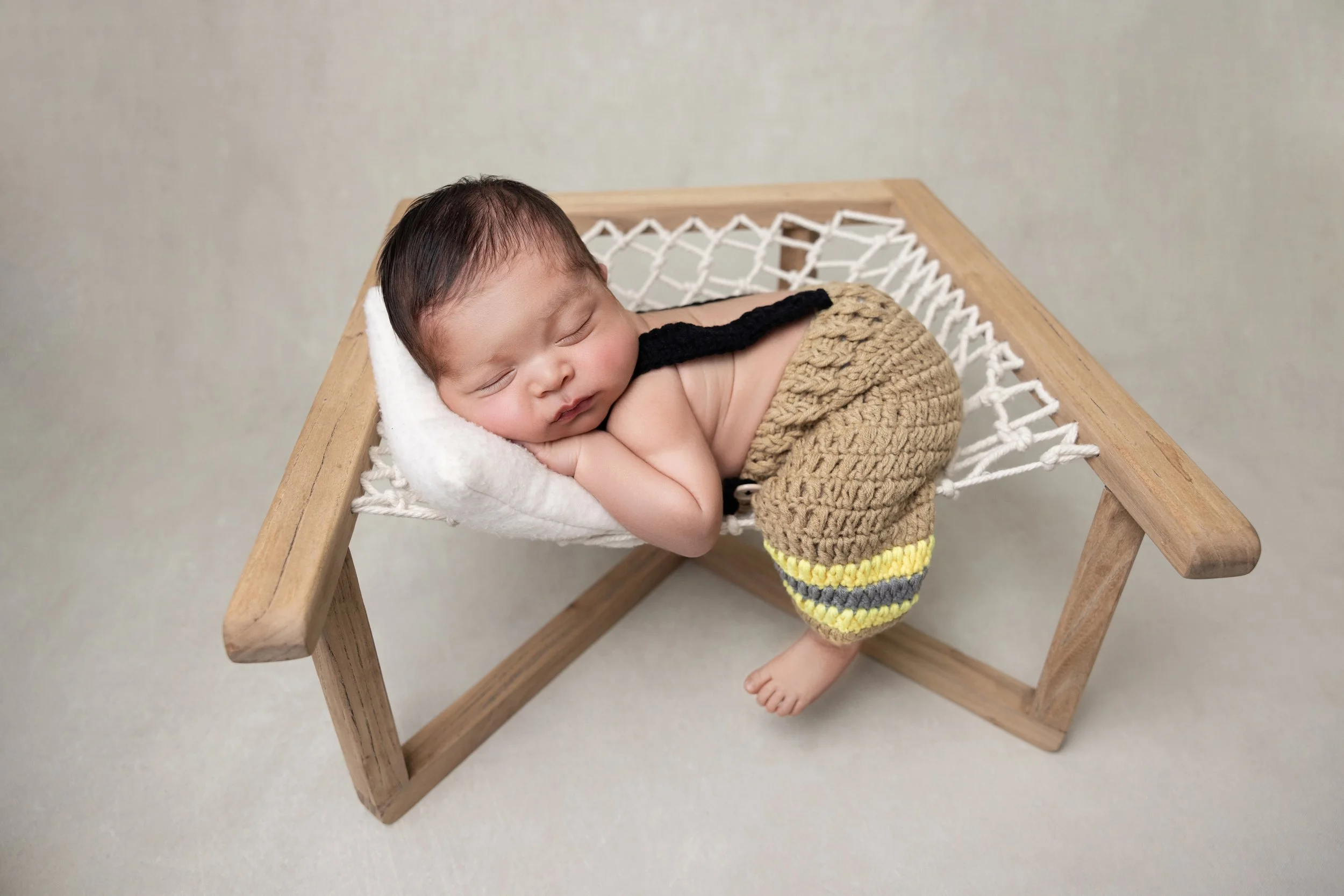 Sleeping baby with dark hair, resting on a small sleep pillow, in a miniature wooden cradle with a crochet mattress, wearing brown knitted pants with yellow and gray stripes.
