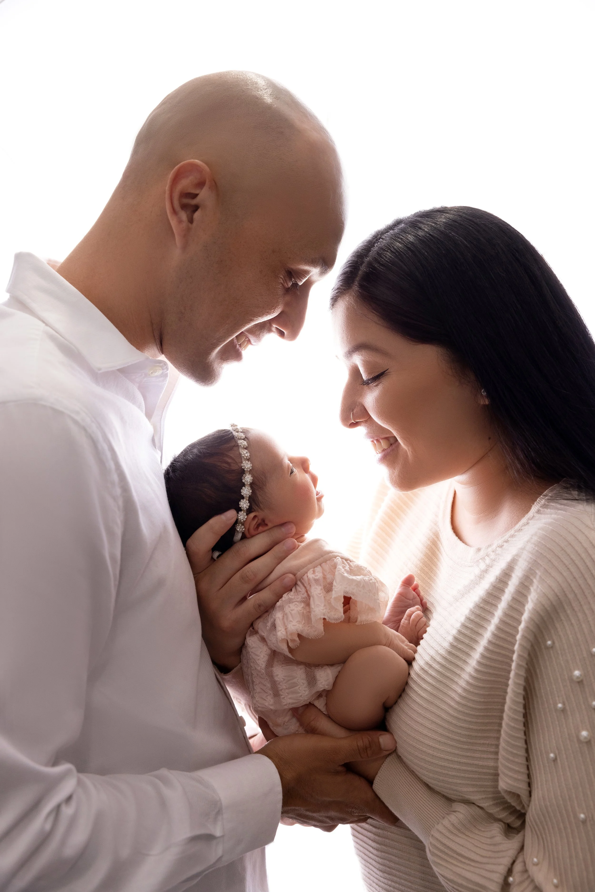 A family of three with a newborn, the father and mother smiling with their foreheads touching, holding the baby between them, against a bright white background.