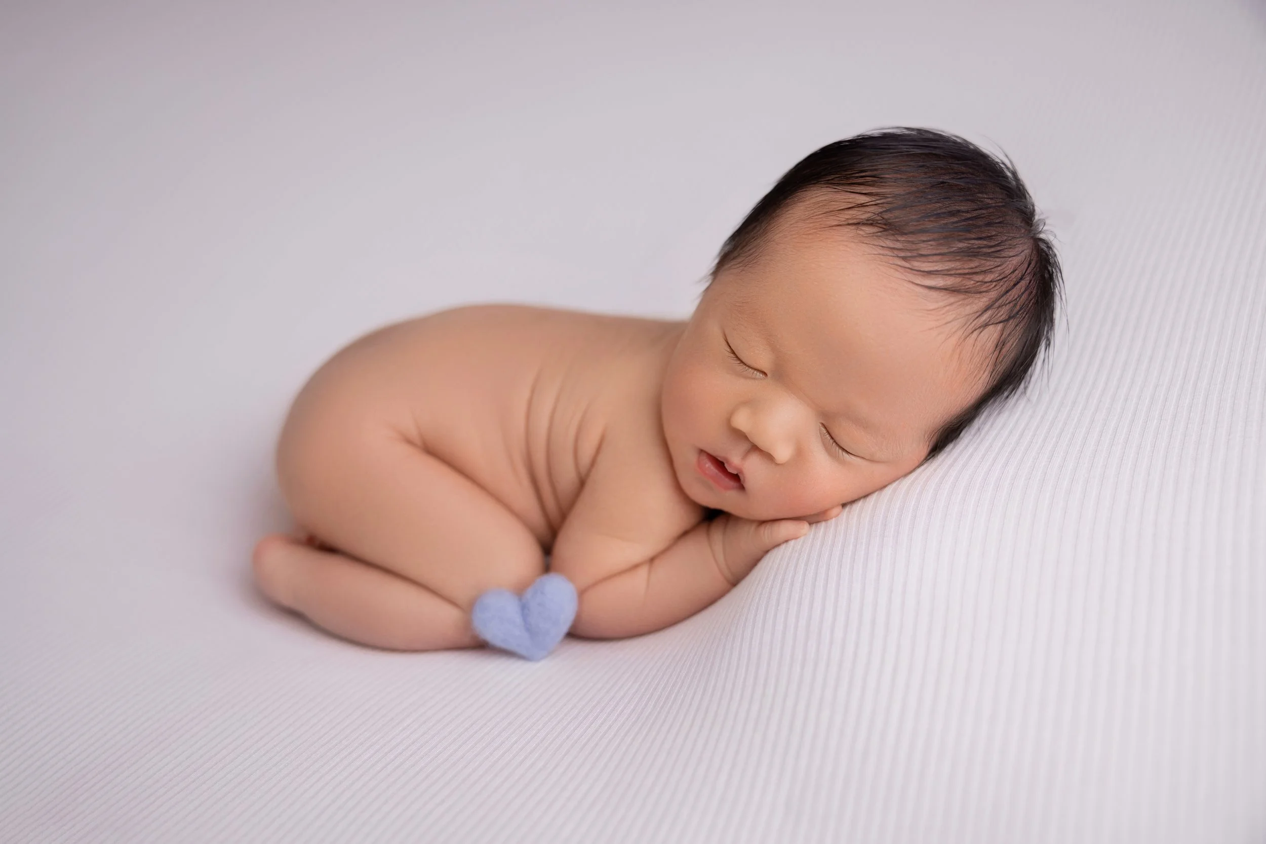 A sleeping baby with short black hair, curled in a fetal position on a white textured surface, wearing a small blue heart-shaped accessory near the feet.