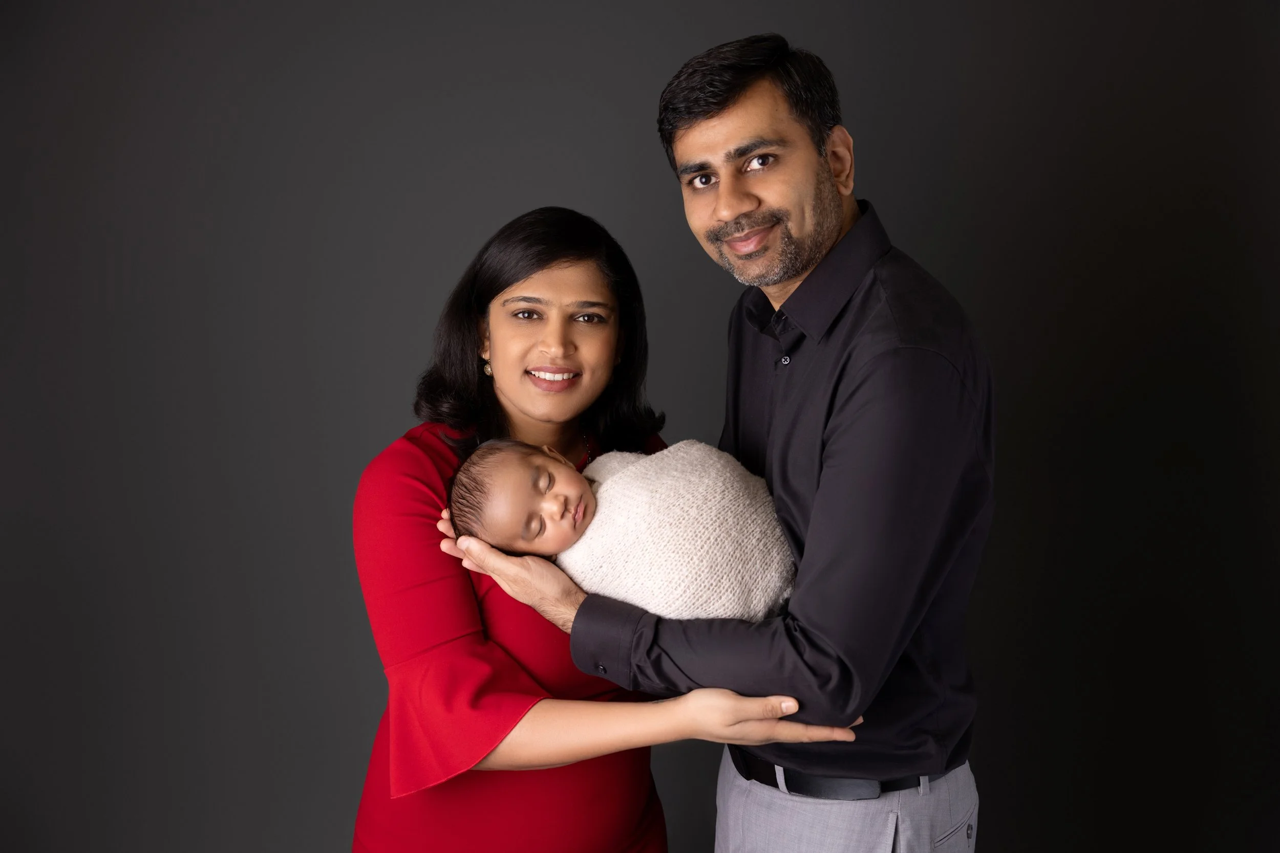 A family portrait with a woman in a red dress, a man in black shirt, and a newborn baby wrapped in a beige blanket, against a dark background.