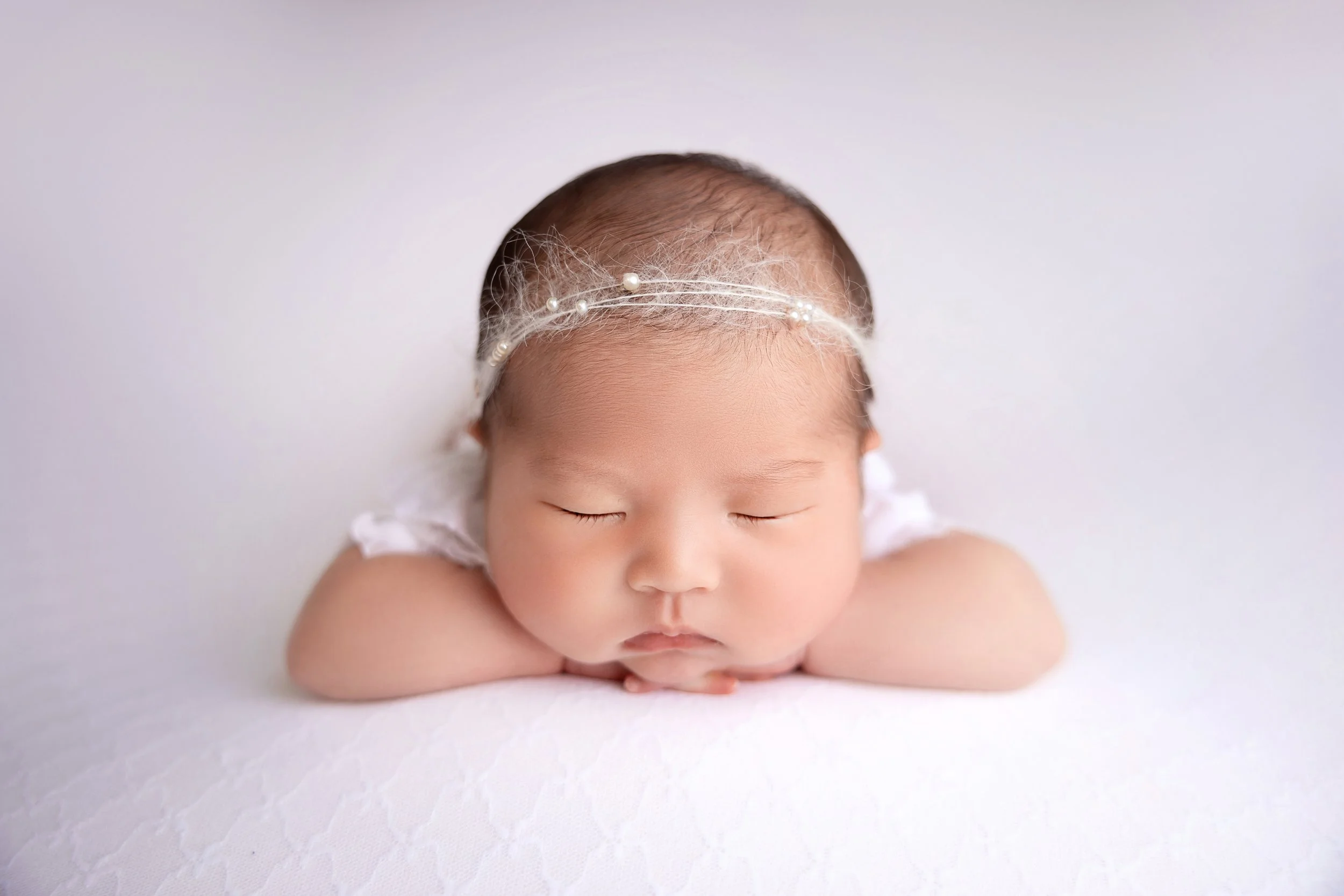 A sleeping newborn baby with a headband of pearls on a white surface