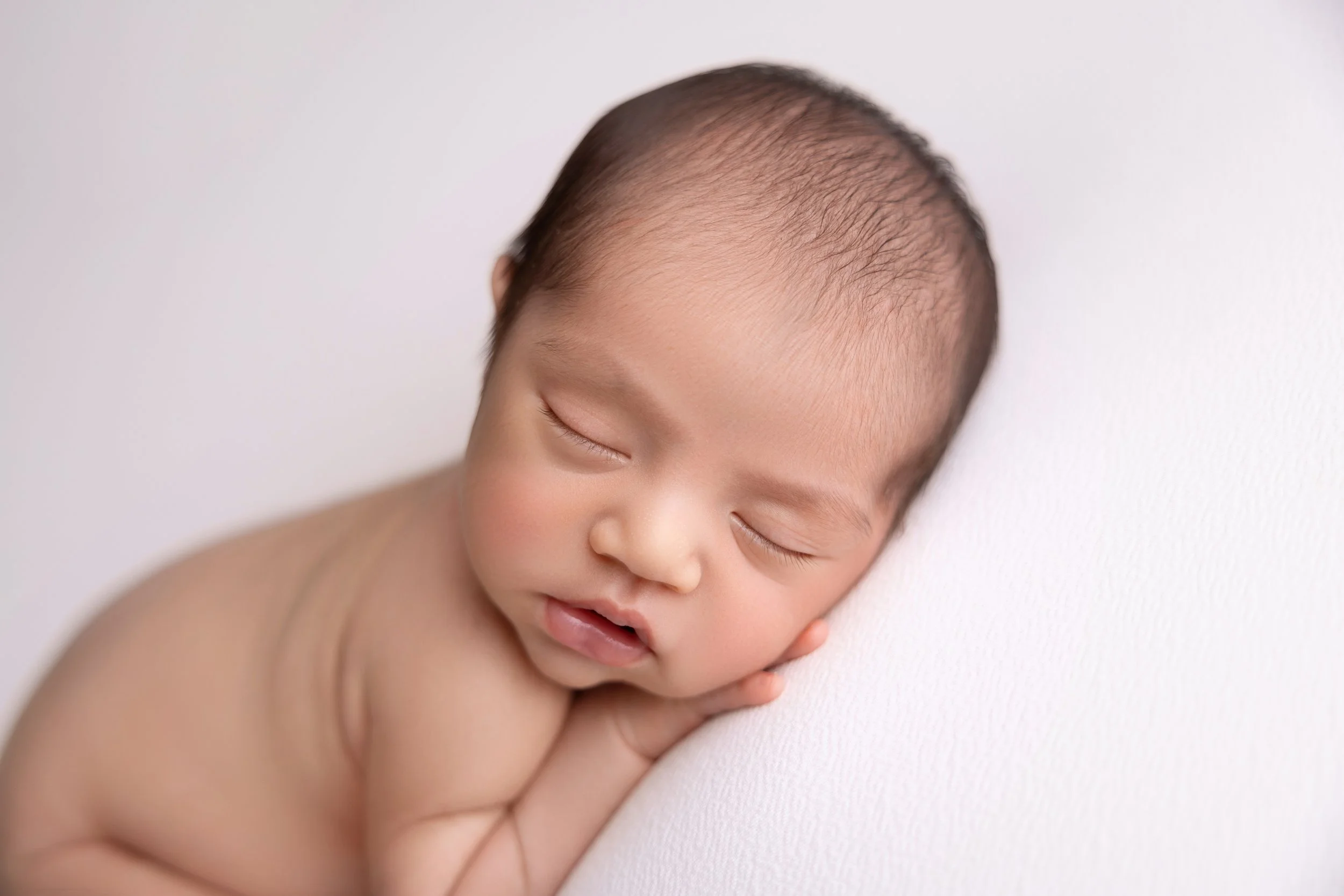Close-up of a sleeping baby with a light skin tone, resting head on a white surface with eyes closed and lips slightly parted.