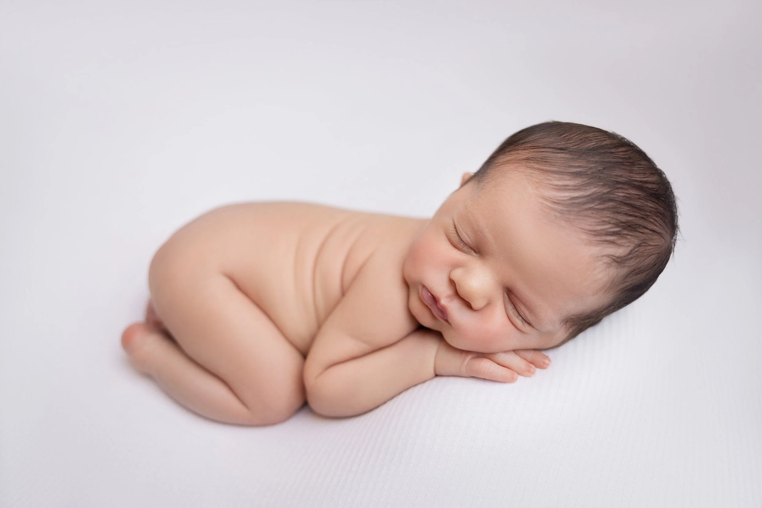 A sleeping newborn baby lying on their side on a white surface, with hands tucked under their face and eyes closed.