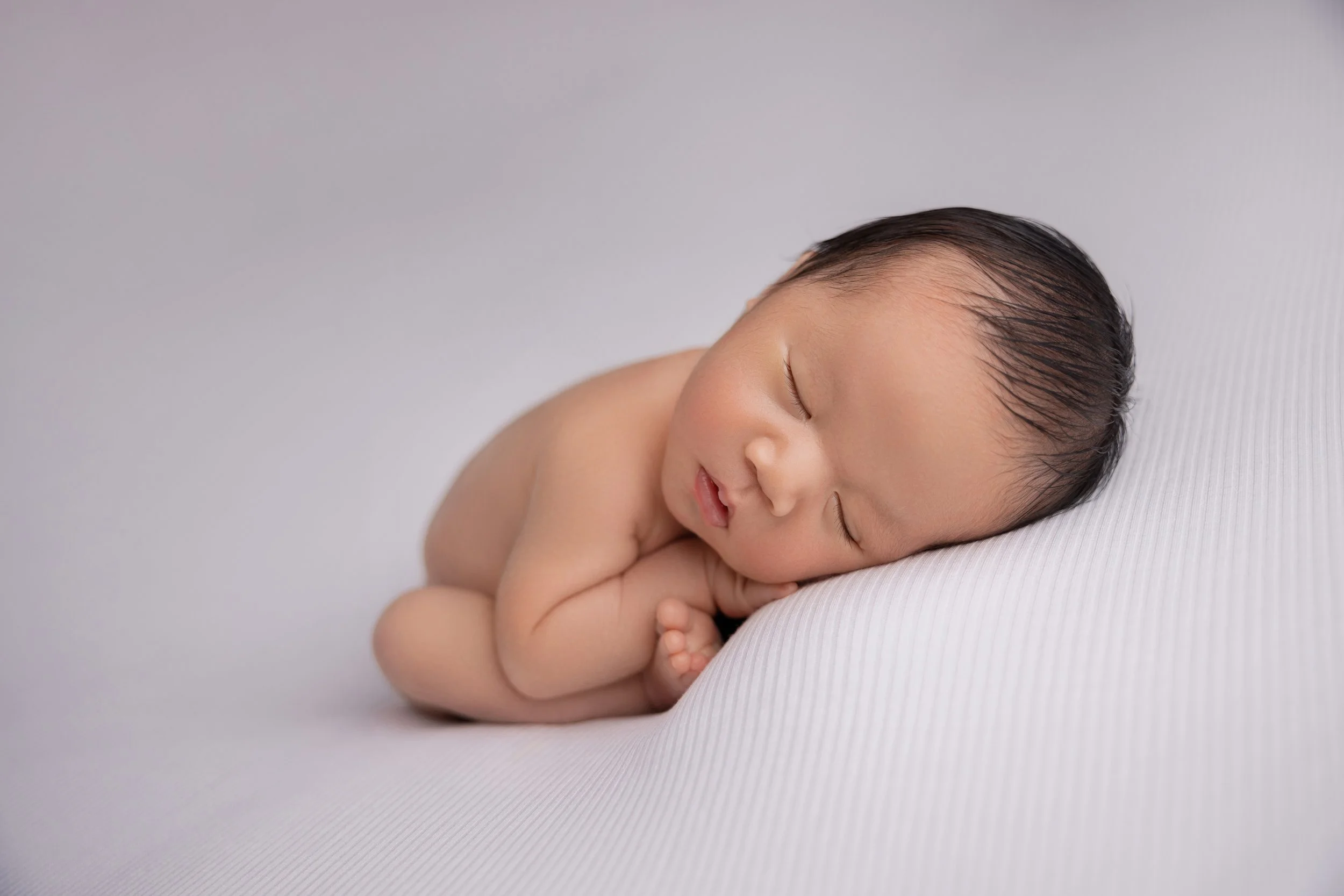 A sleeping newborn baby curled up with the head resting on a white surface