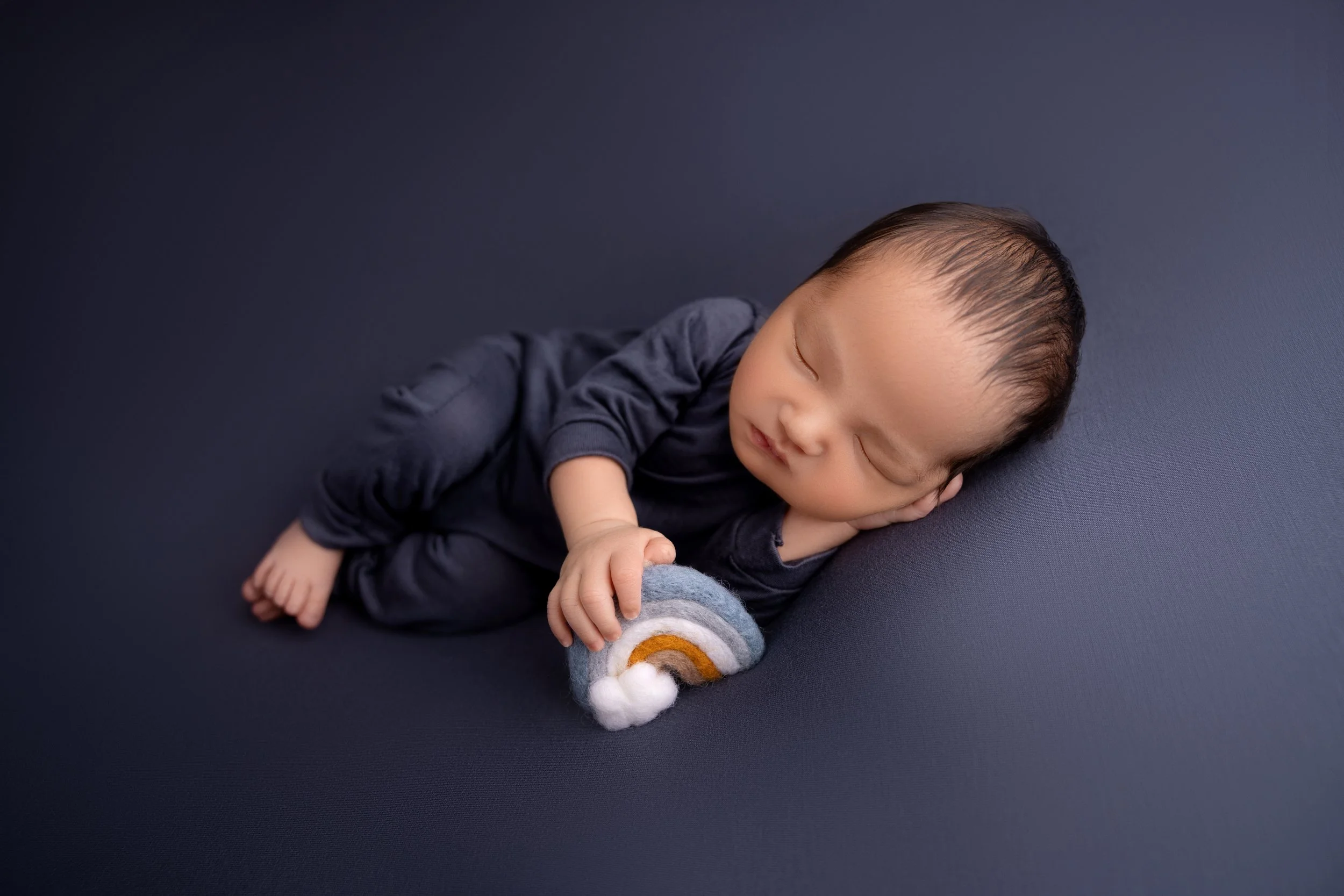 A sleeping baby lying on a dark blue surface, holding a small rainbow and cloud plush toy.