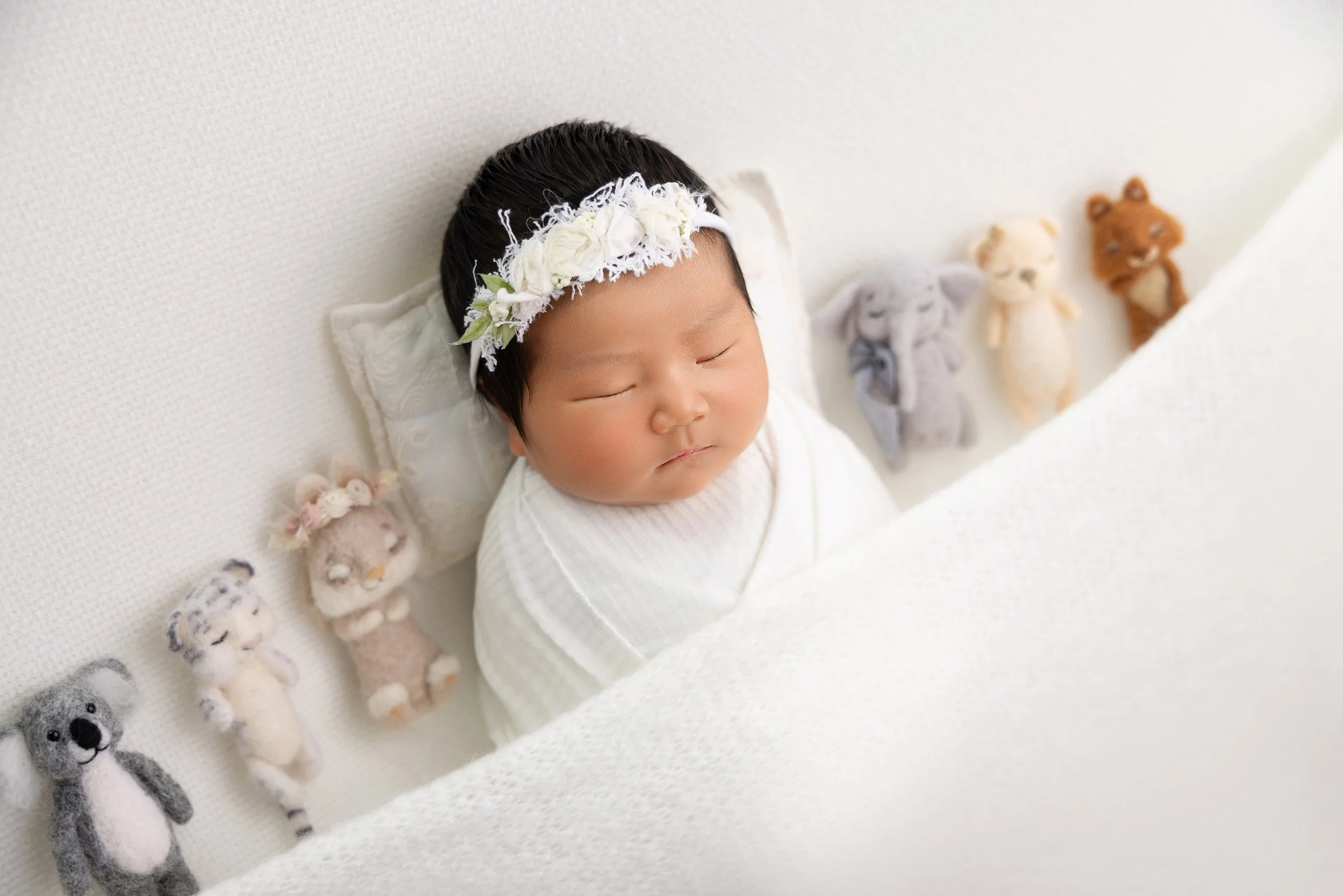A sleeping Asian baby girl with a floral headband, wrapped in a white blanket, lying on a white textured surface surrounded by small plush animal toys.