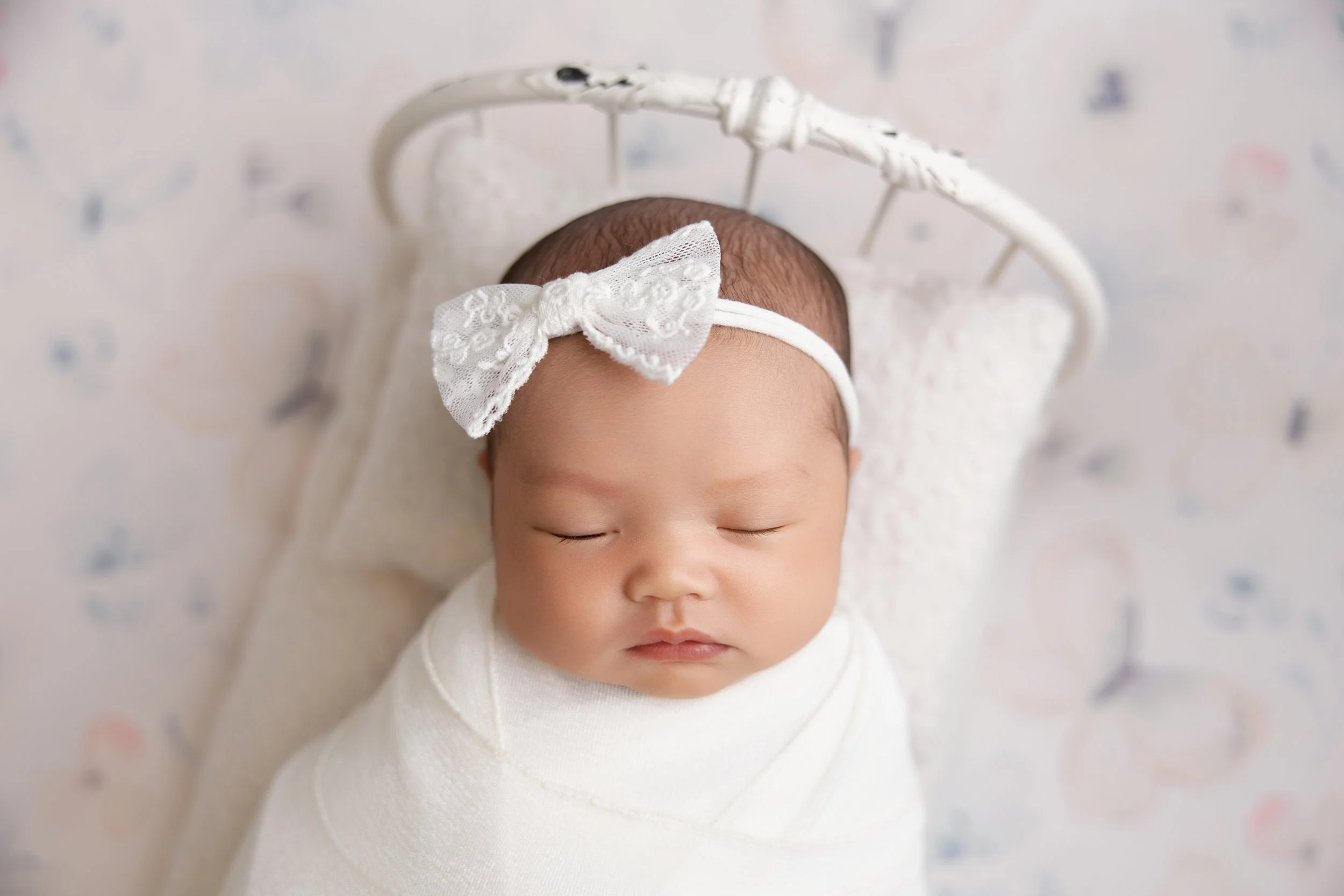 Close-up of a sleeping newborn baby wrapped in a white blanket, with a lace bow headband on her head, lying on a soft pillow.