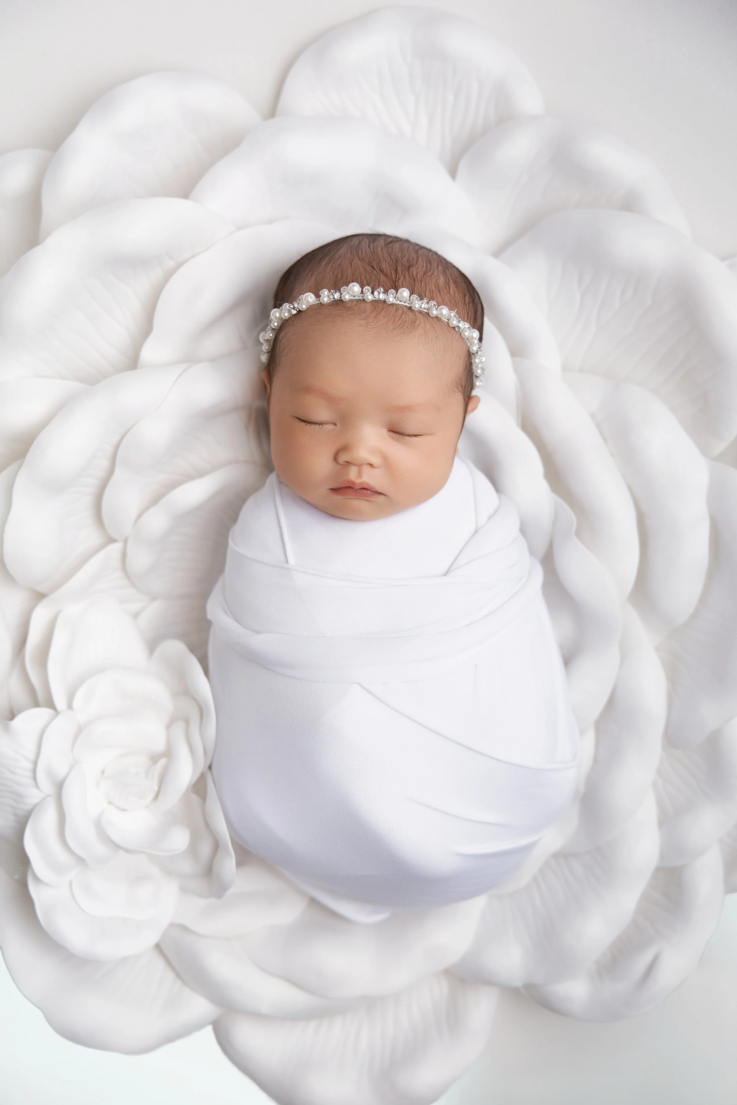 A sleeping baby wrapped in a white blanket, wearing a pearl and bead headband, lying on a decorative white floral prop.