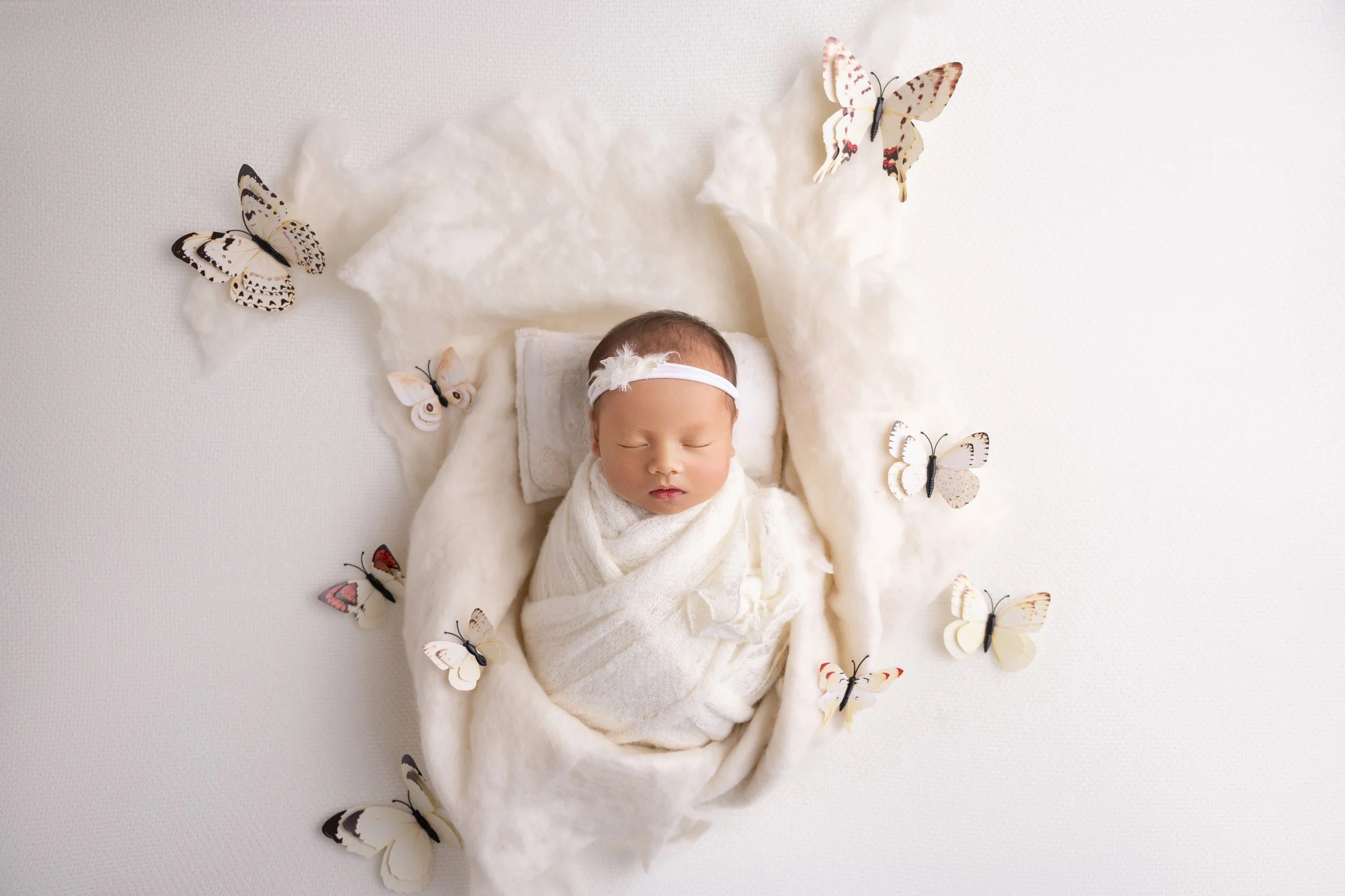 A sleeping baby wrapped in a white blanket, lying on a soft platform surrounded by artificial butterflies on a white background.