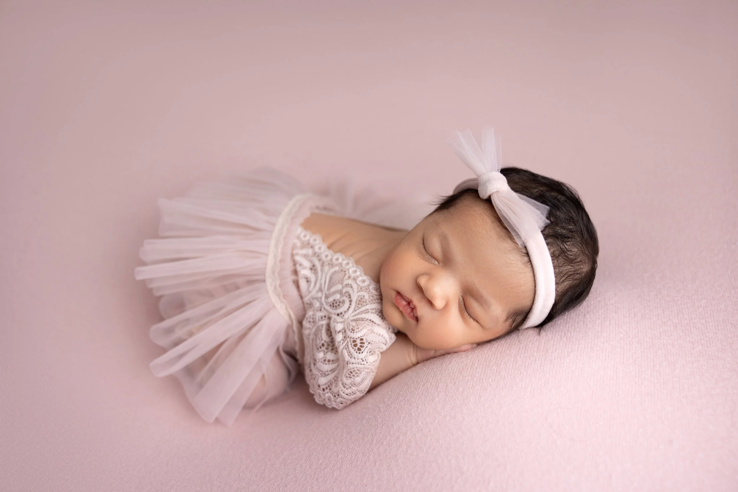 A sleeping baby girl with dark hair, wearing a pink tulle dress and a matching headband with a bow, lying on a pink surface.
