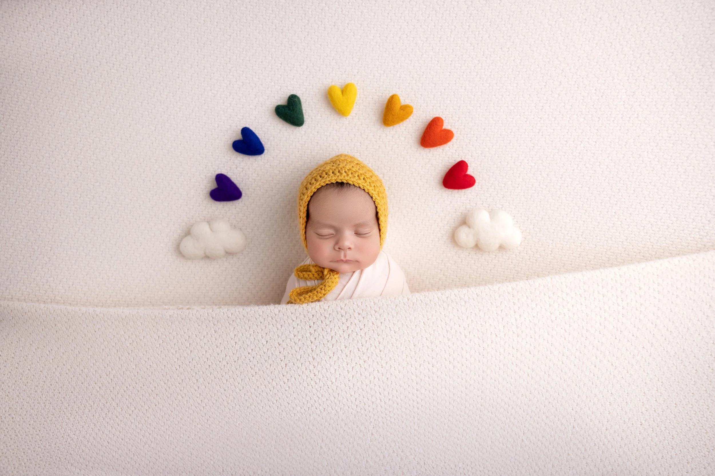 A sleeping baby wearing a yellow knit hat with a chin strap, lying on a cream-colored textured blanket. Above the baby, there are rainbow-colored felt hearts arranged in an arc and small white felt clouds on either side.
