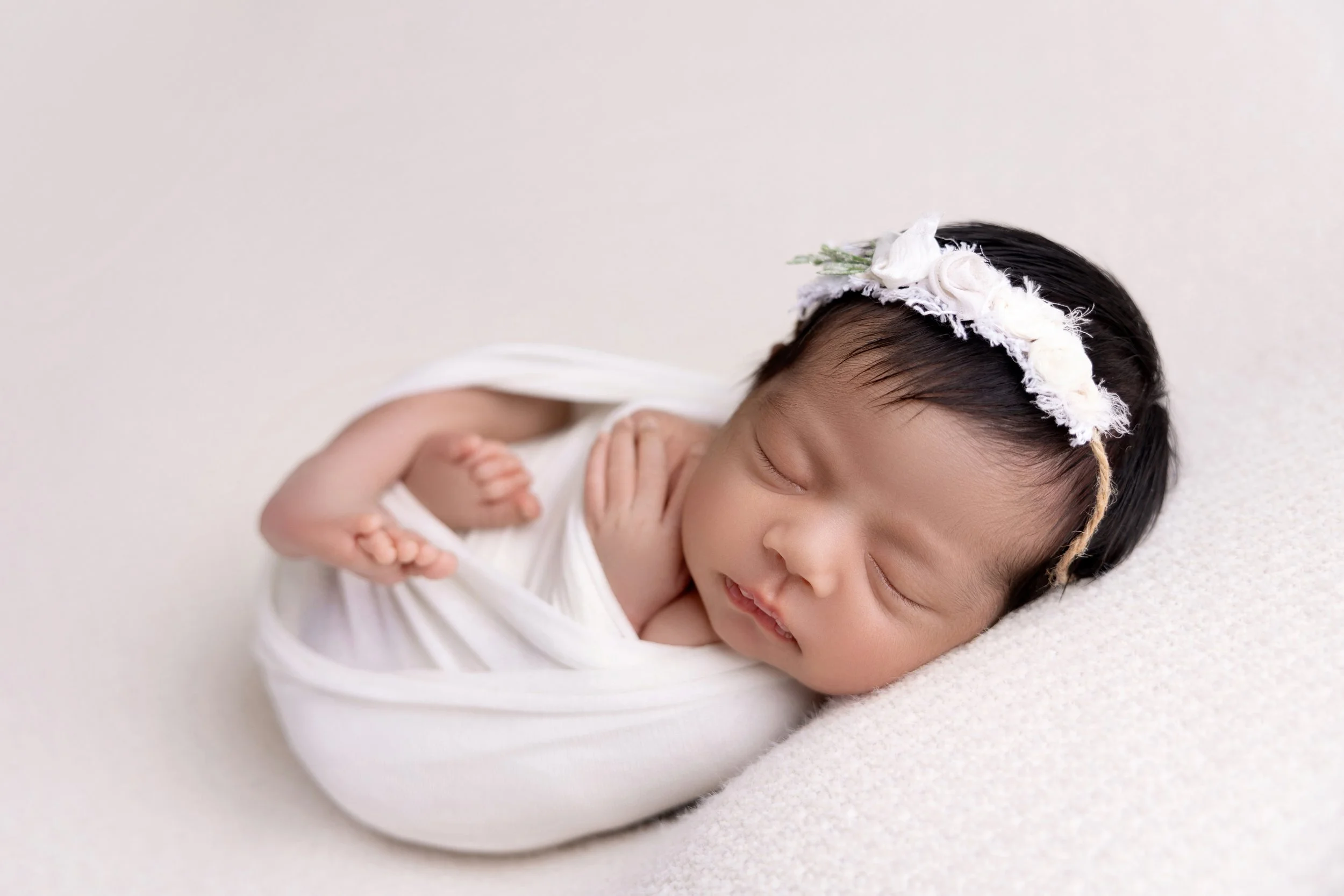 A sleeping baby with a floral headband, wrapped in a white cloth, lying on a soft surface.