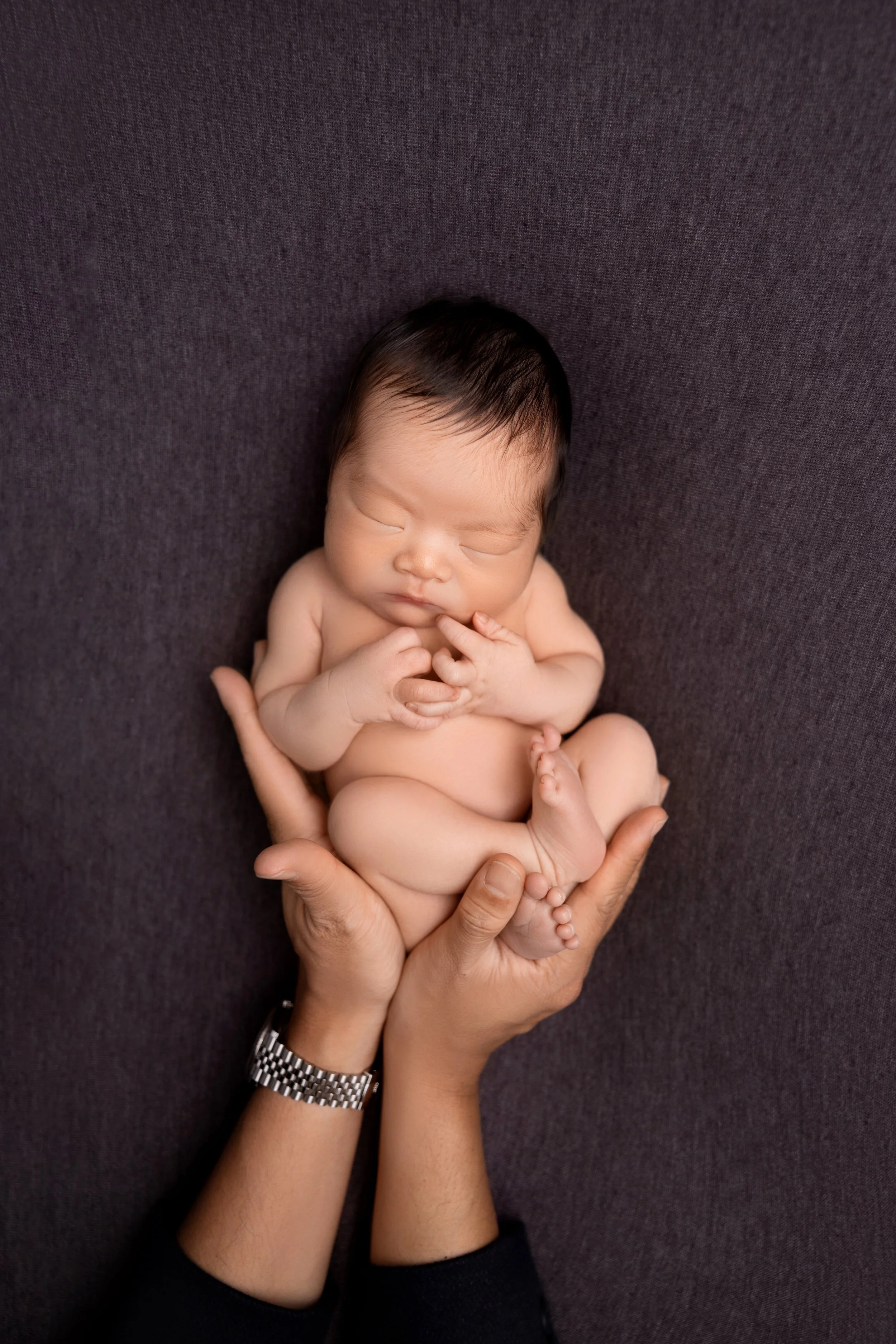 A newborn baby with dark hair sleeping peacefully, held gently in an adult's hands against a dark gray background.
