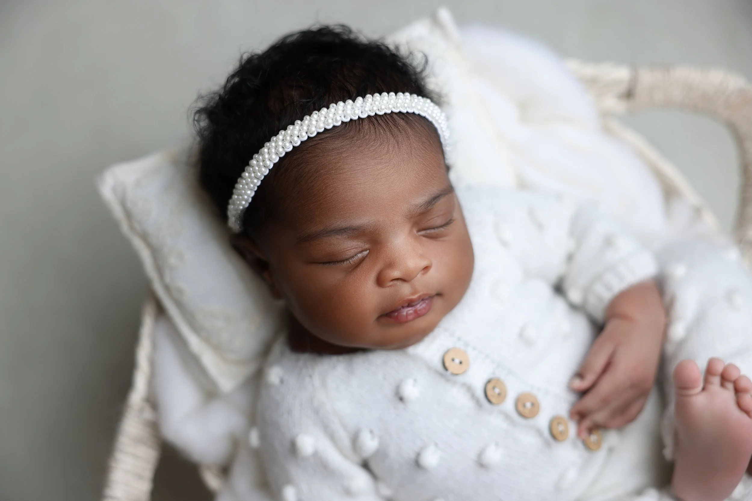 A sleeping baby wearing a pearl headband and a white knitted sweater, lying on a soft pillow with a neutral background.