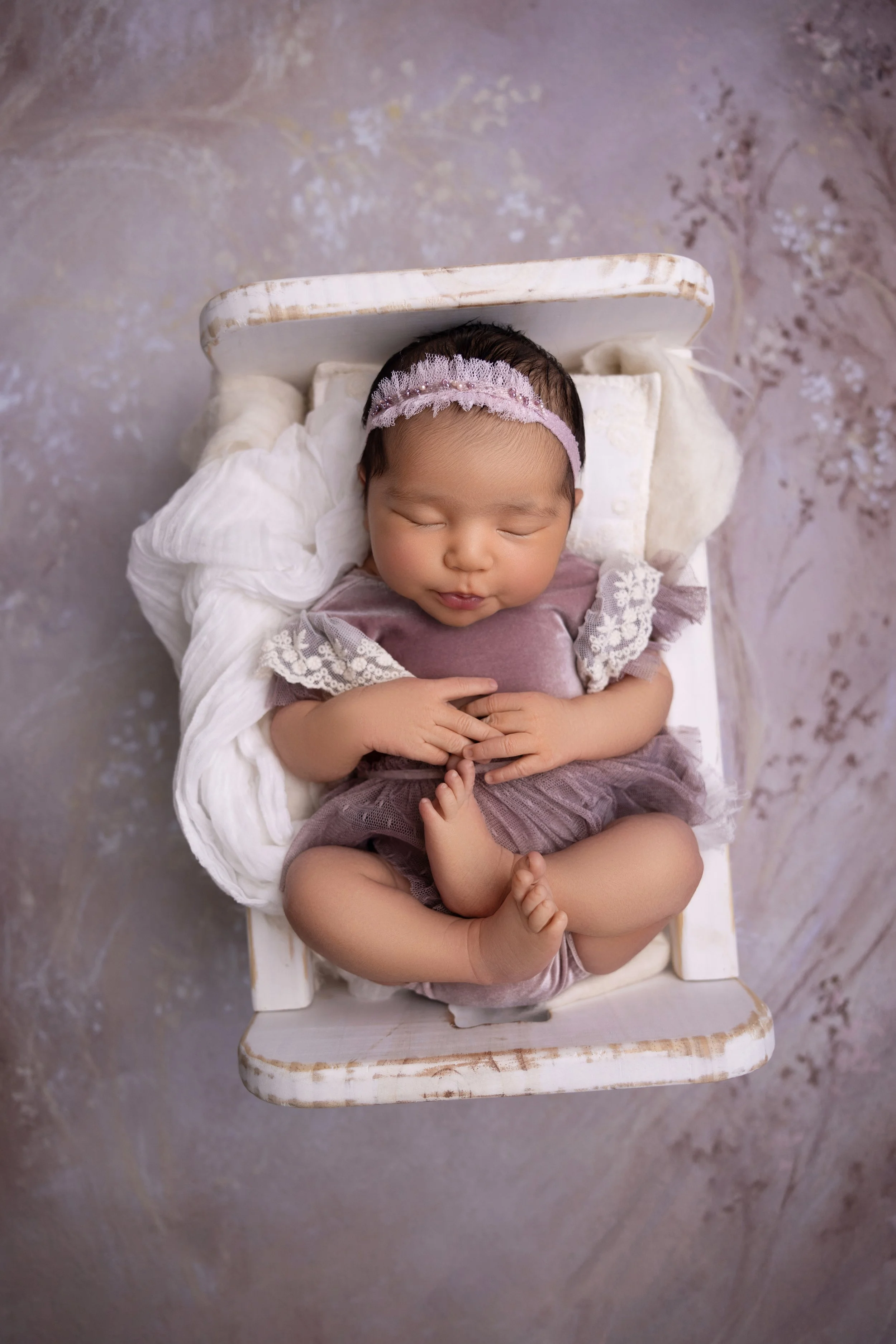 A sleep baby girl lying on a small white wooden bed, wearing a pink headband and a purple lace dress, on a textured surface background.