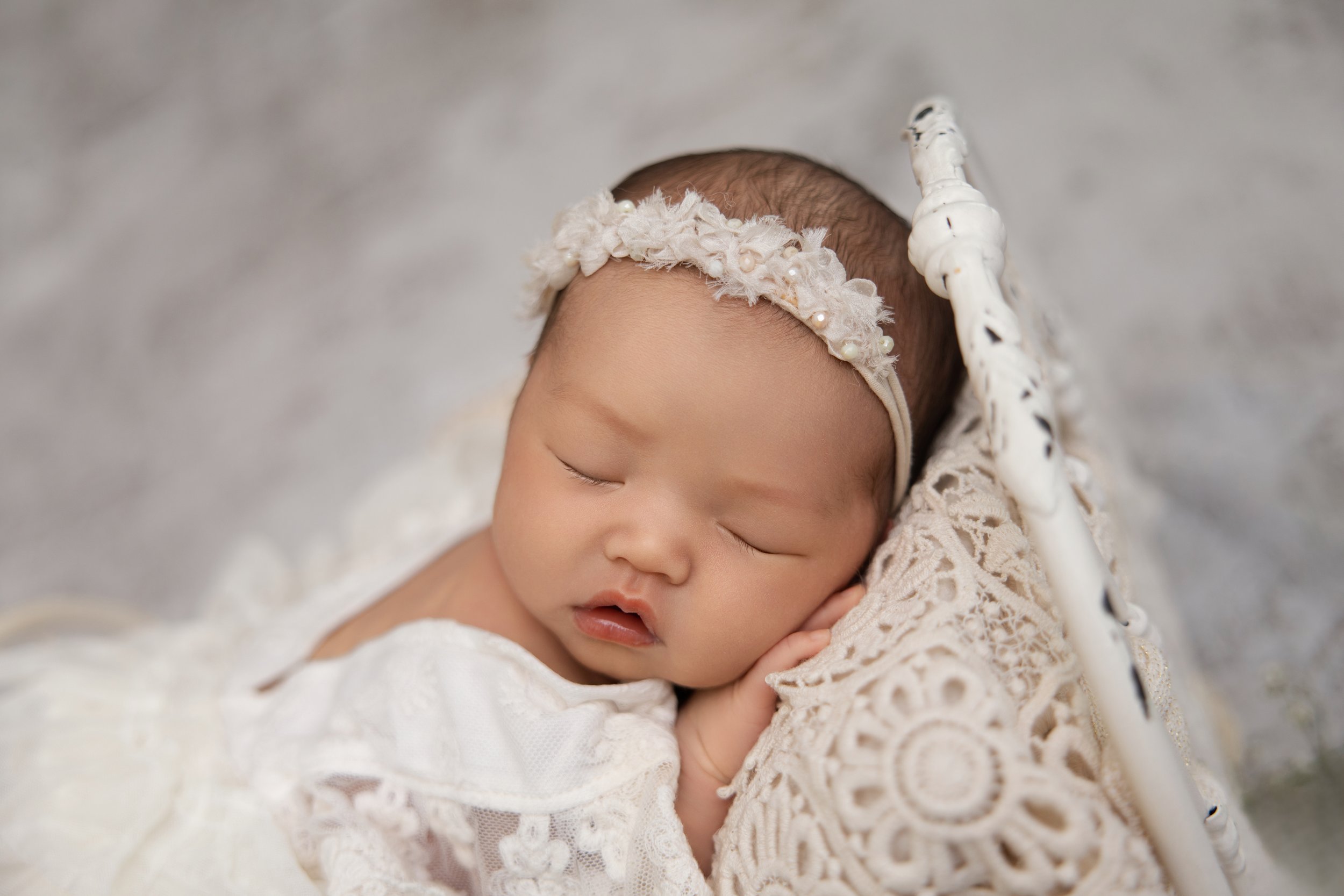 A sleeping baby with a floral headband, resting on a lace blanket in a cradle.