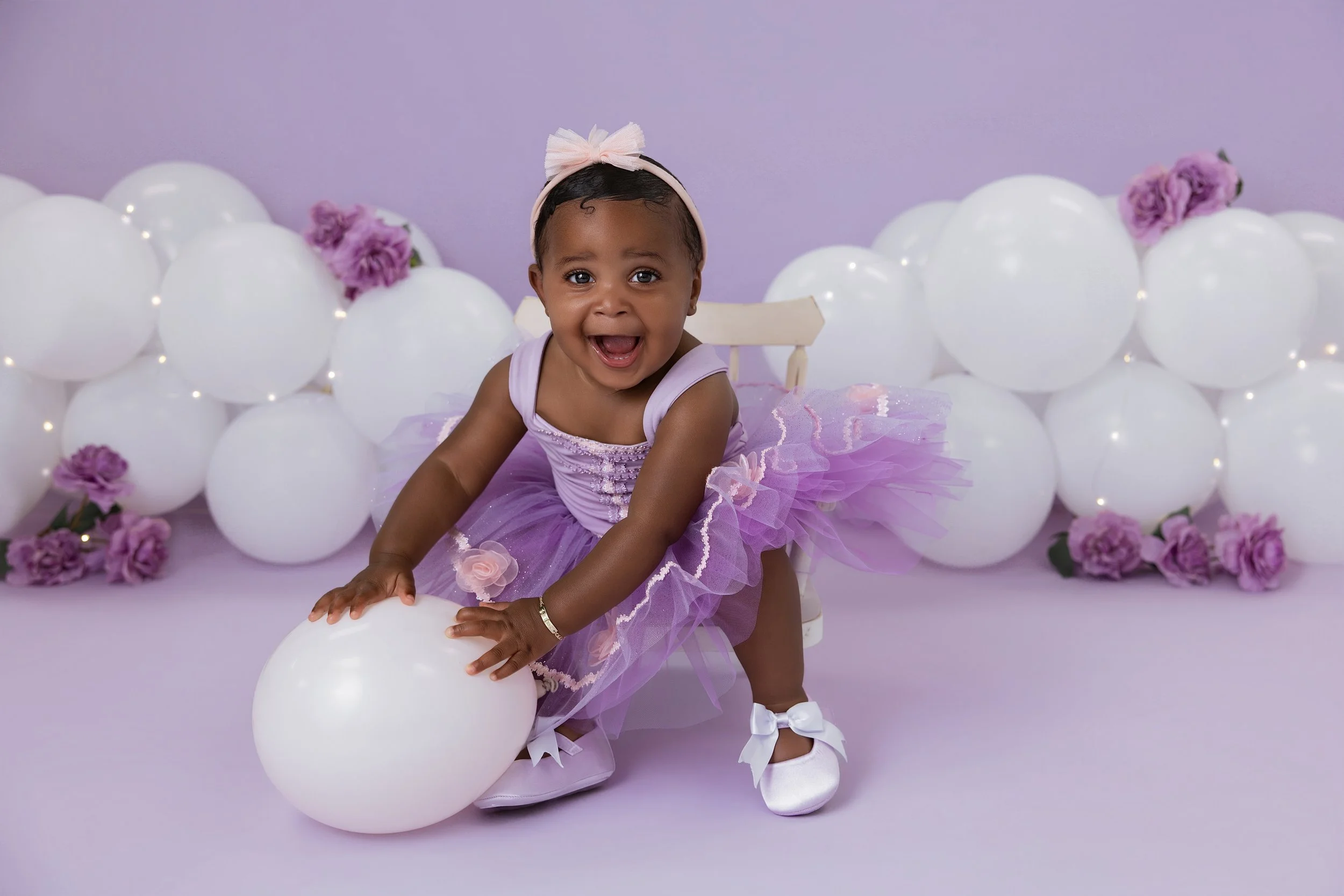 Young girl celebrating her first birthday, sitting on the floor with a cake, surrounded by flowers and decorative items.