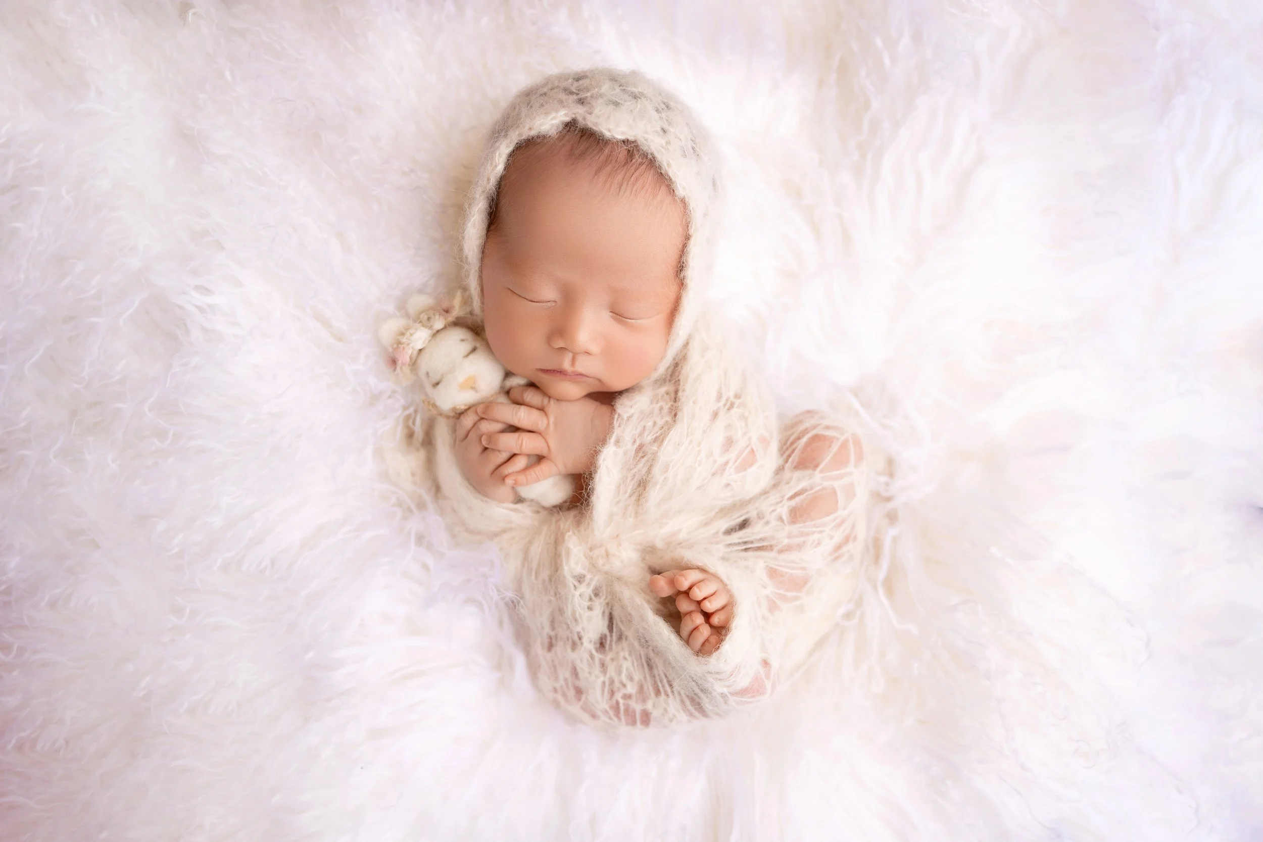 A sleeping newborn baby wrapped in a soft, cream-colored knit blanket with a matching hood, holding a small plush toy, lying on a fluffy white surface.
