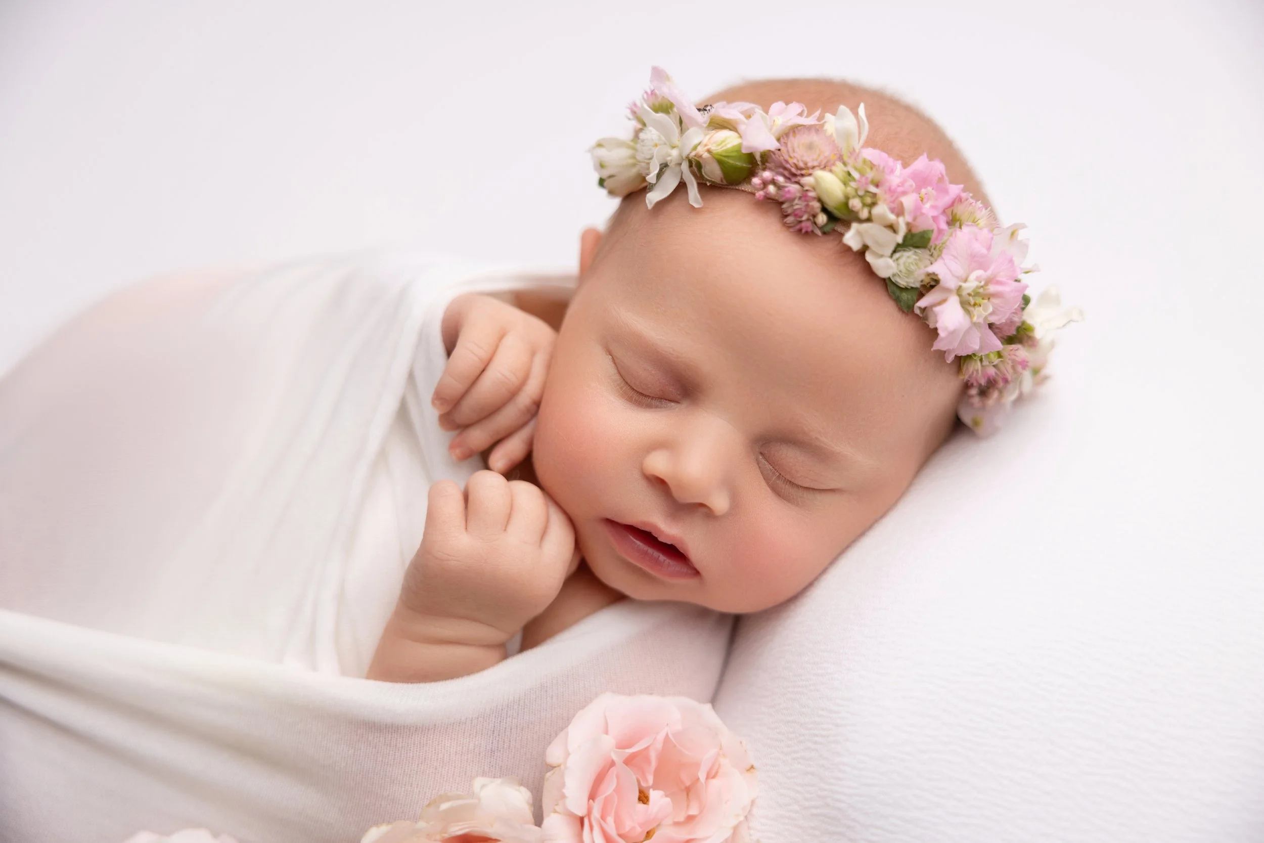 A newborn baby with a floral crown sleeping peacefully on a white pillow, with pink roses nearby.