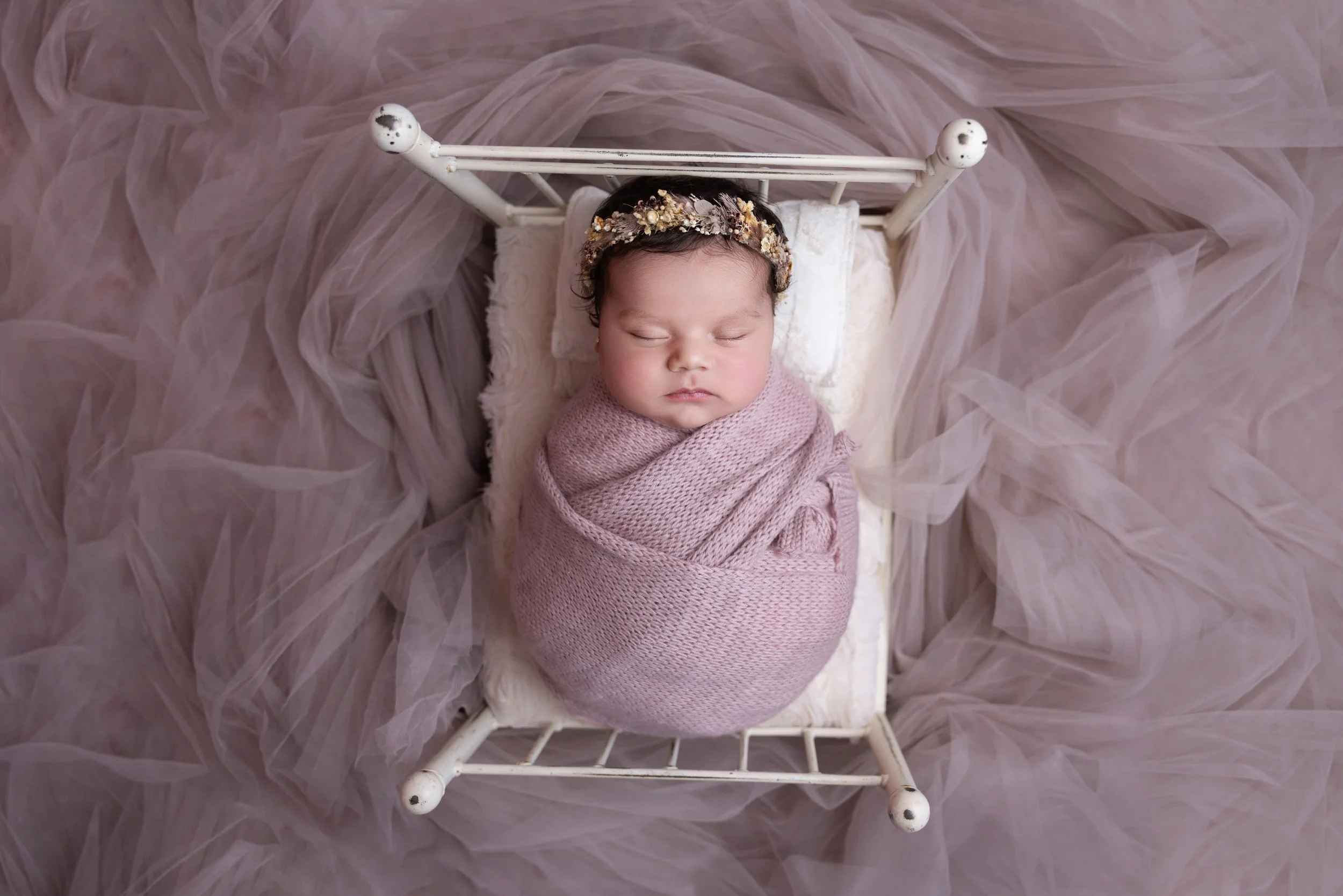A newborn baby girl sleeping on a small white bed, wrapped in a pink knitted blanket, wearing a floral headband, surrounded by pink tulle fabric.