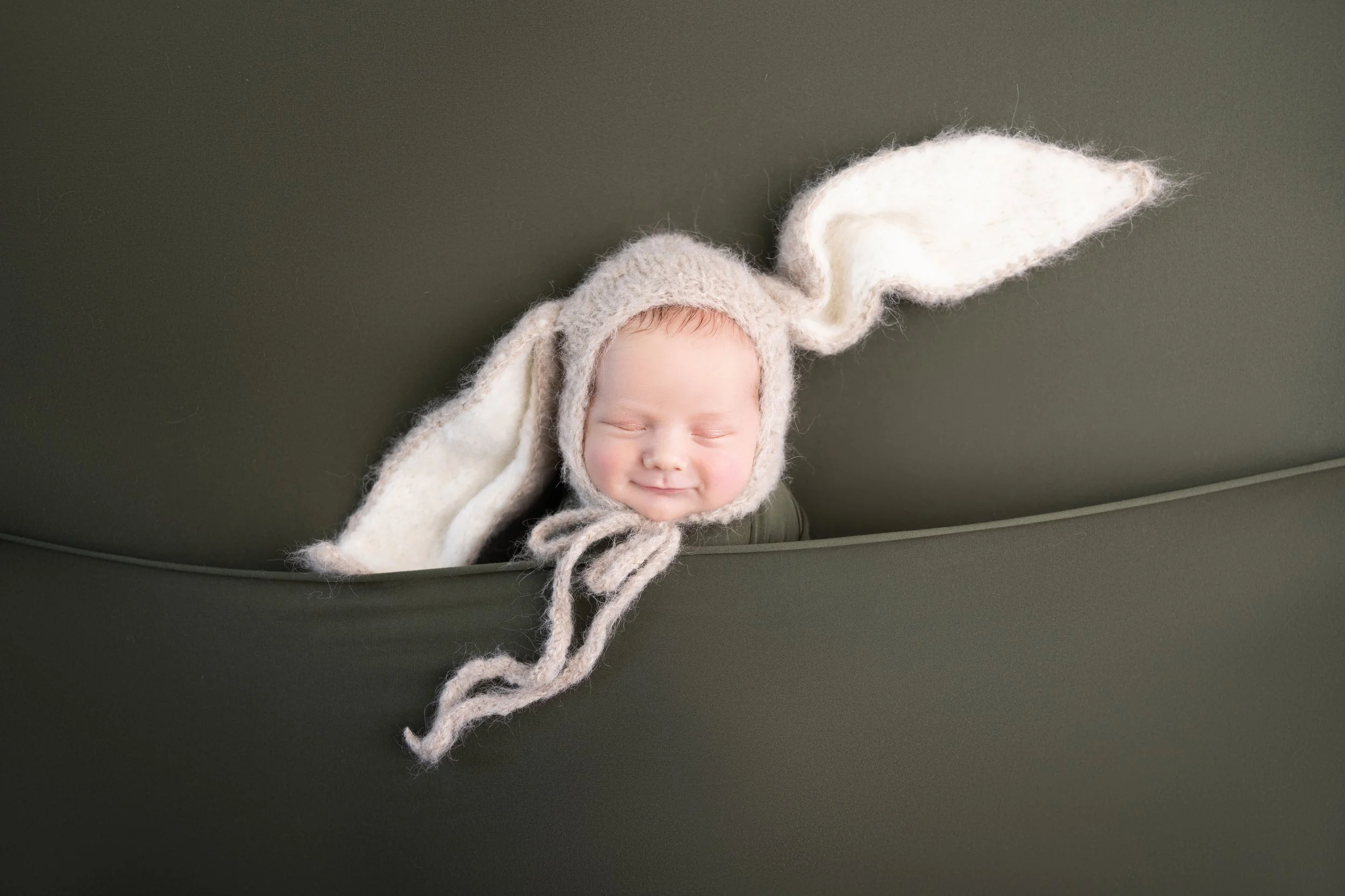 A smiling baby wearing a knitted bunny hat with long ears, nestled in a green basket.