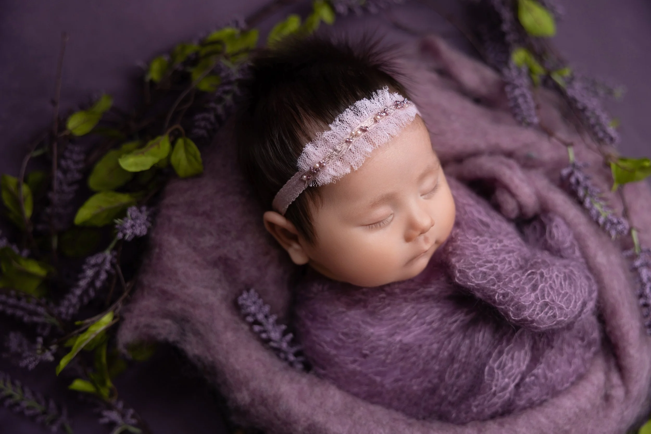 A sleeping baby girl with a pink lace headband, nestled in purple knitted blankets, surrounded by purple flowers and green leaves.